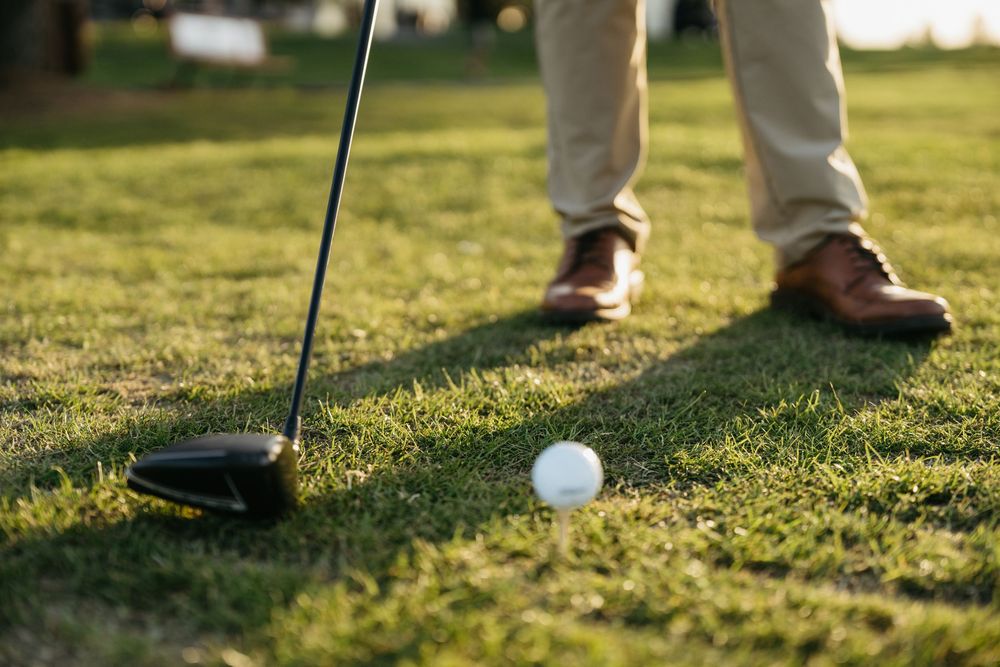 Golfer lines up a driver shot on lush green fairway, wearing khaki pants and brown leather shoes.