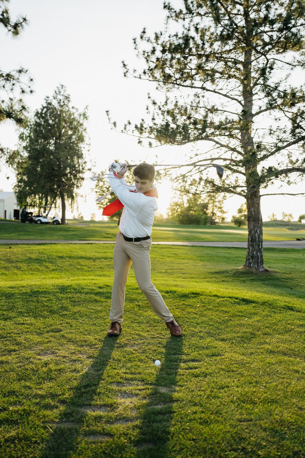 Golfer completing a full swing on a sunny golf course with lush green fairway and trees in background.