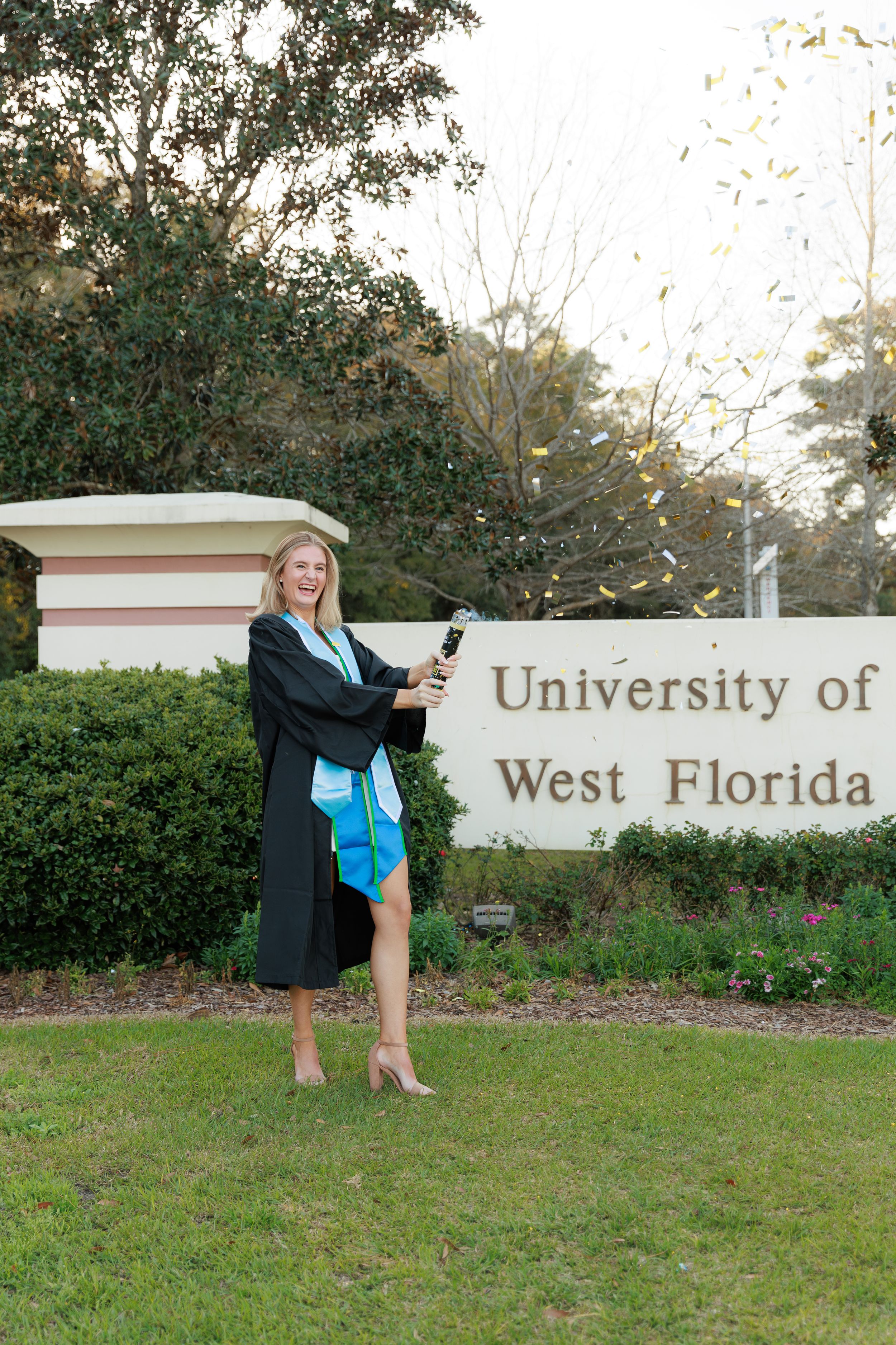 A graduate in cap and gown poses with diploma in front of the University of West Florida entrance sign.