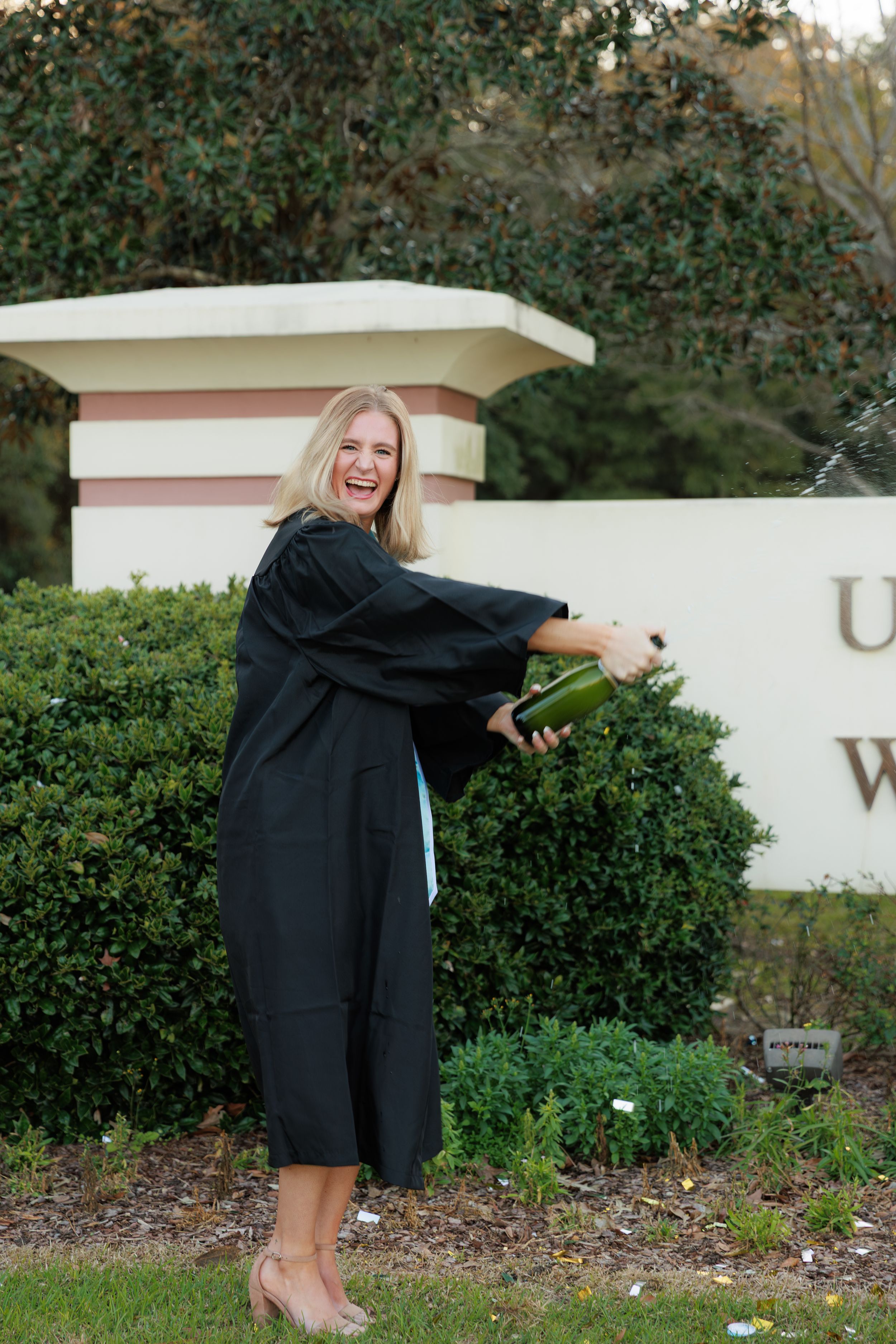 Graduate in black cap and gown poses playfully by university entrance sign during graduation photo session.
