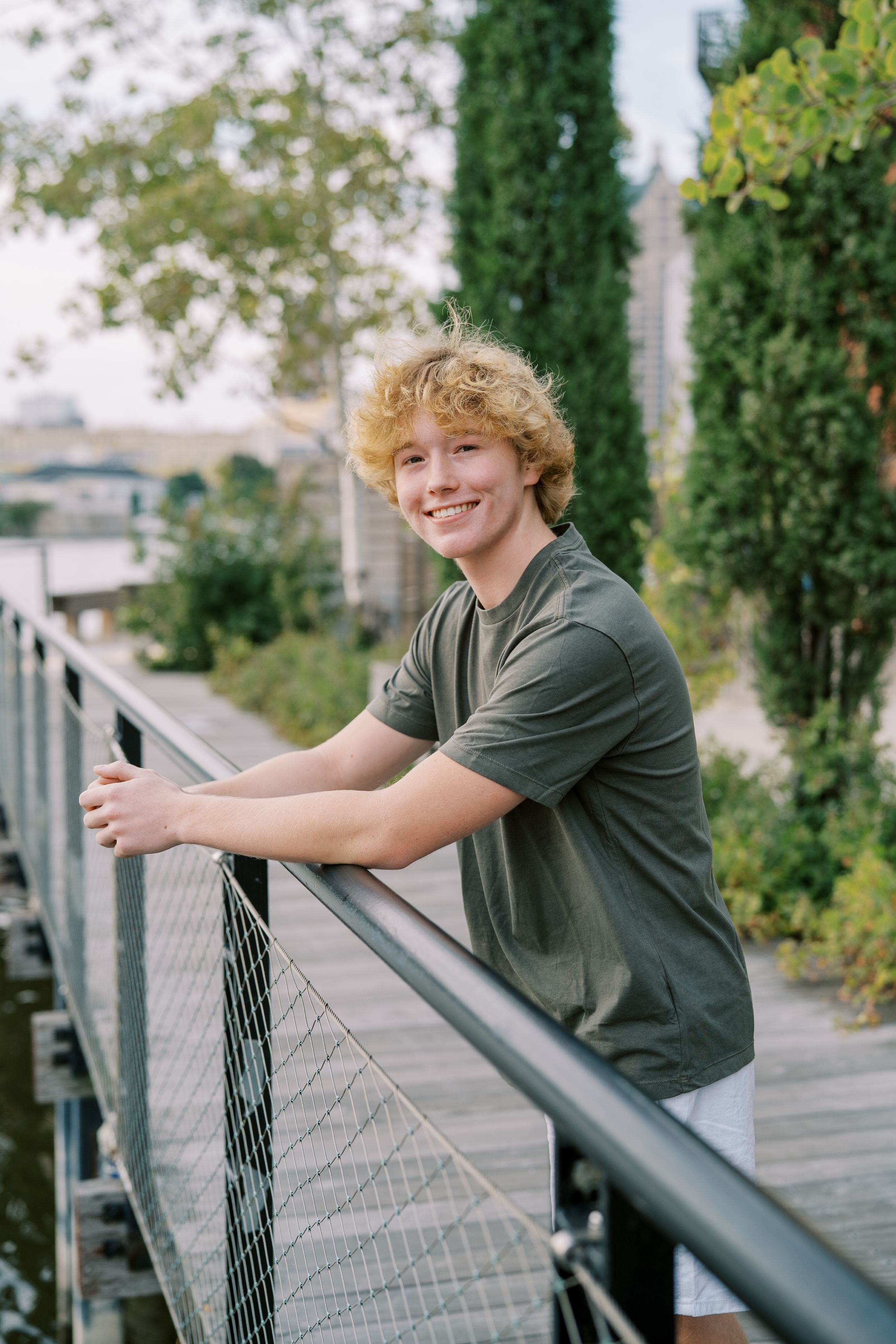 Relaxed pose by railing wearing olive t-shirt with greenery in background.