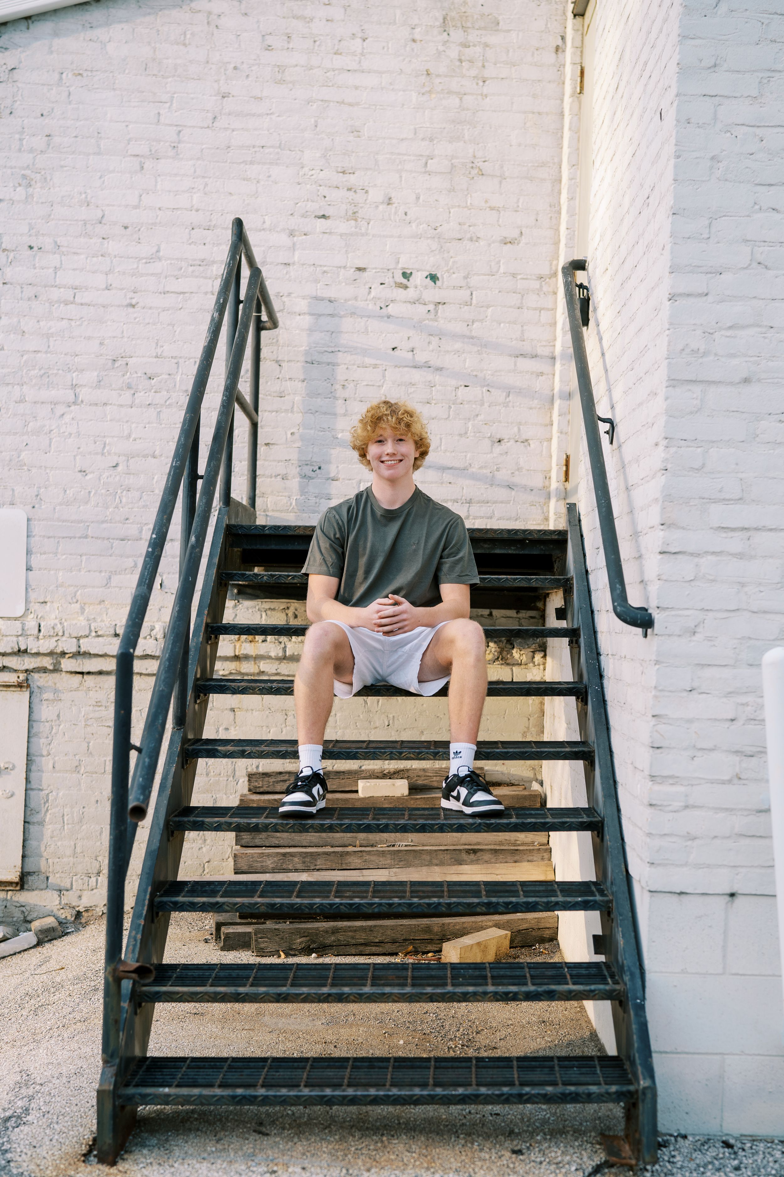 Casual portrait seated on metal staircase against white brick wall.