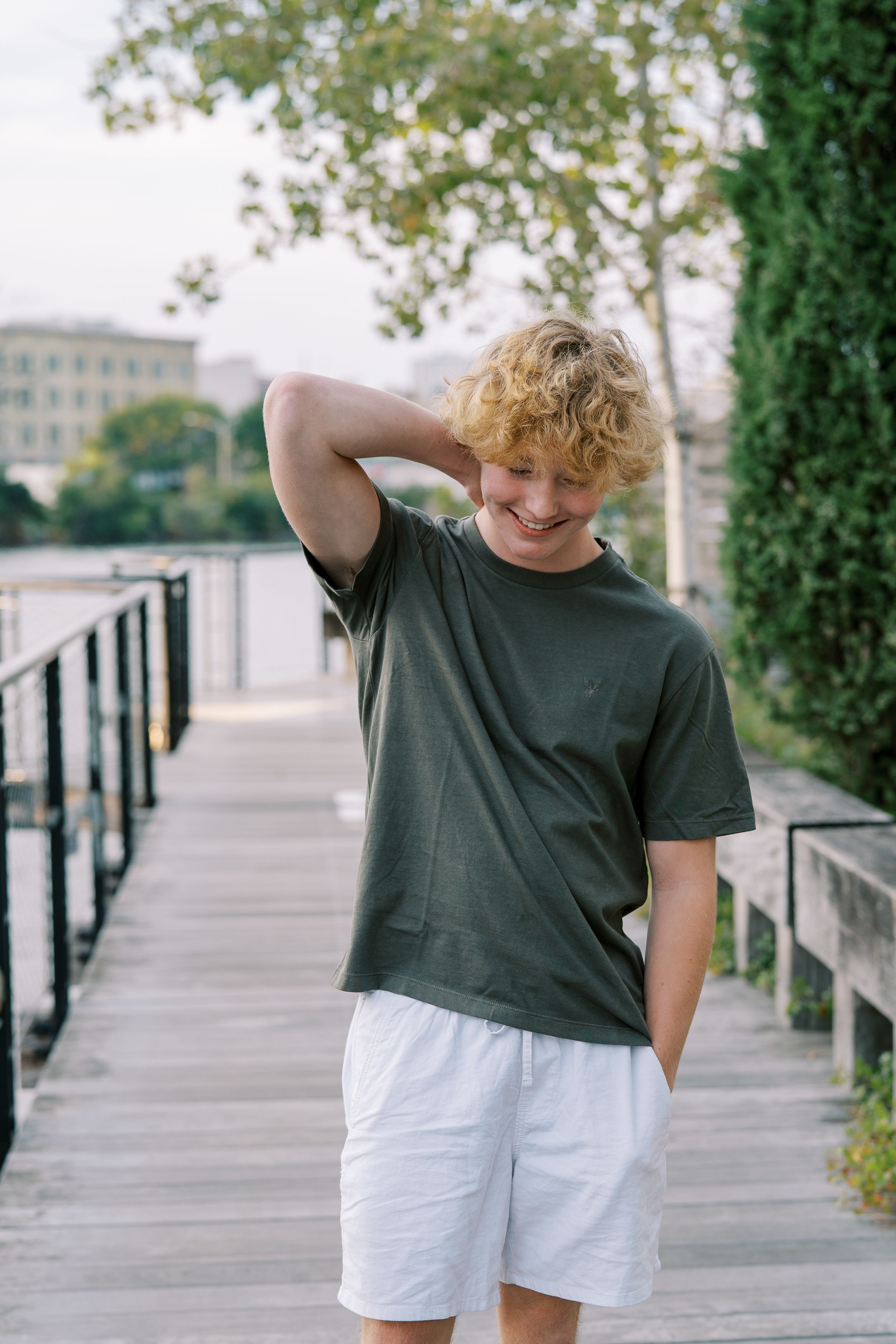 Person in dark shirt and white shorts poses casually on a wooden boardwalk with trees in background.