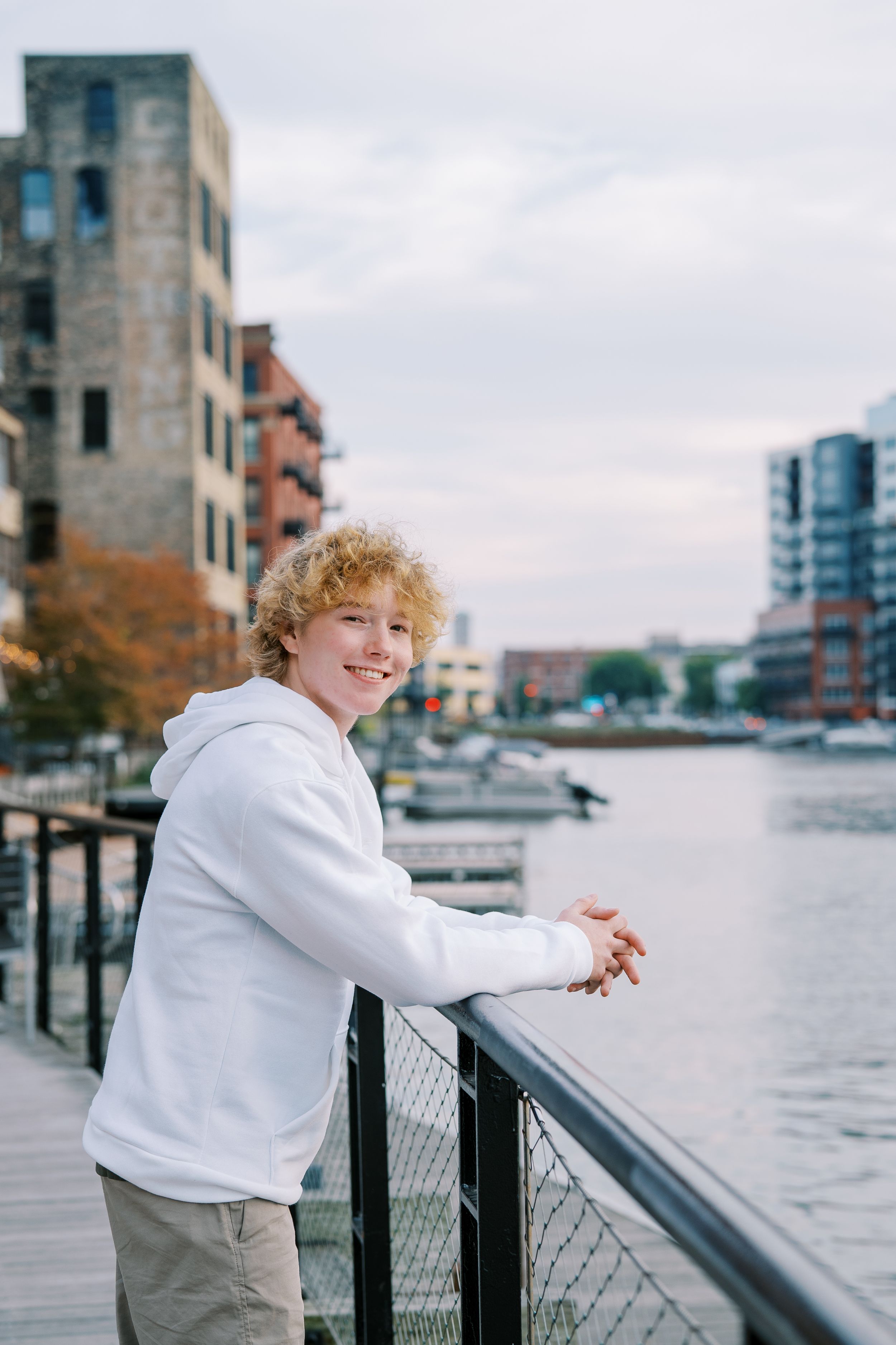 Person in white sweater leans on railing overlooking waterfront with urban buildings in background.