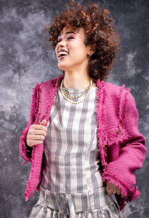 Girl in a pink blazer with a beige plaid dress on, laughing  with hands on hips looking up and to the right, shot from low angle