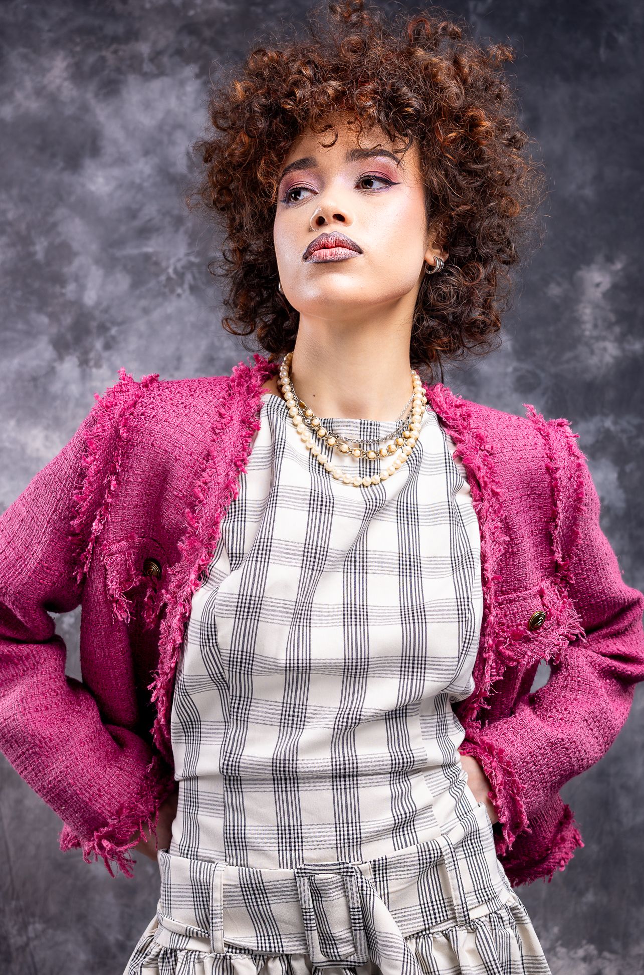 Girl in a pink blazer with a beige plaid dress on, with hands on hips looking up and to the right, shot from low angle