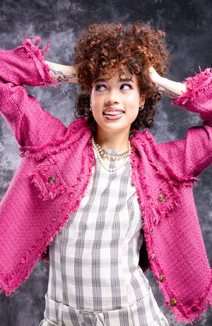 Girl in a plaid dress and blazer, hands in her hair, looking to the left with tongue partially out, smiling