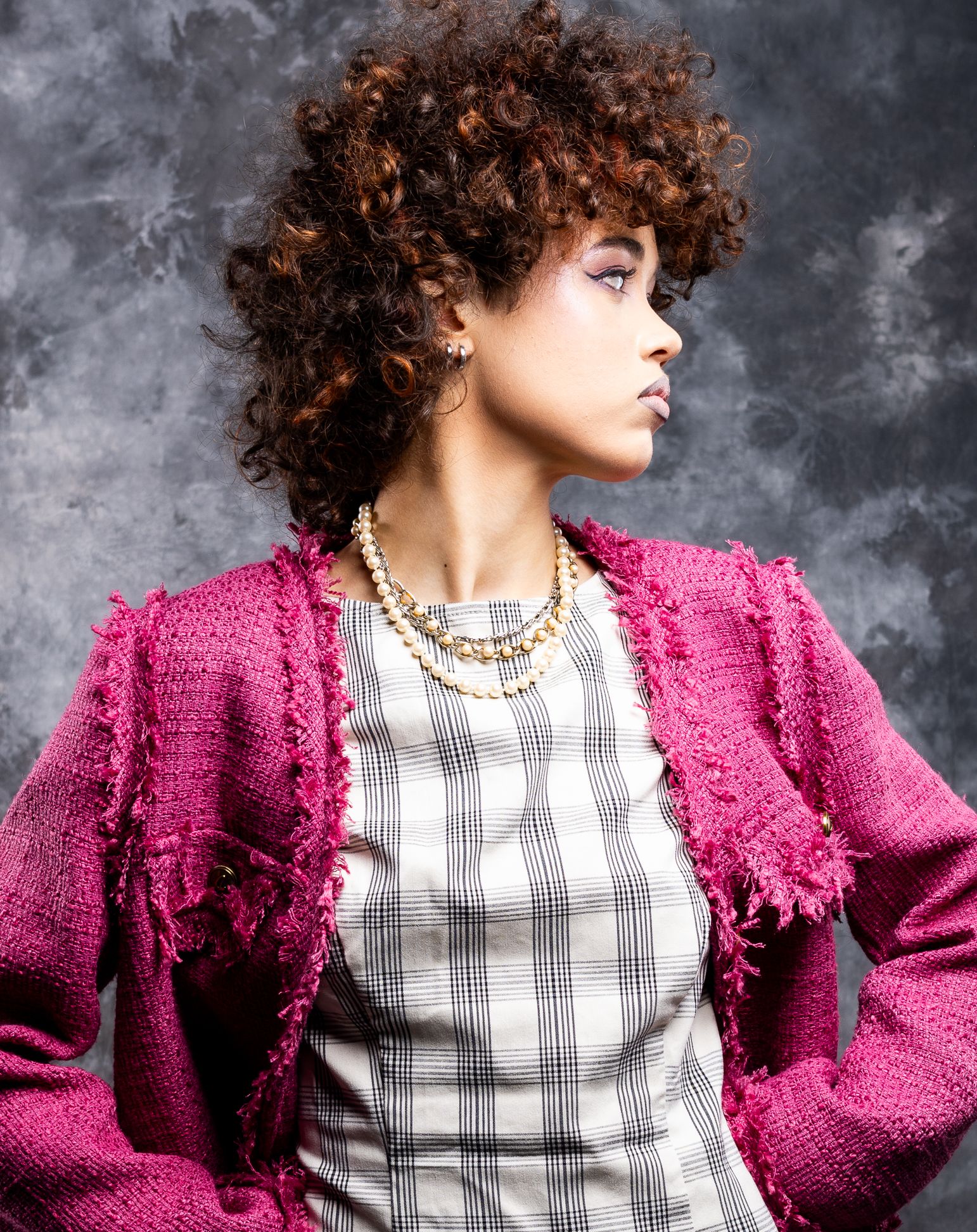 Side profile image of a girl in a pink blazer, beige plaid dress, hands on her hips, looking right