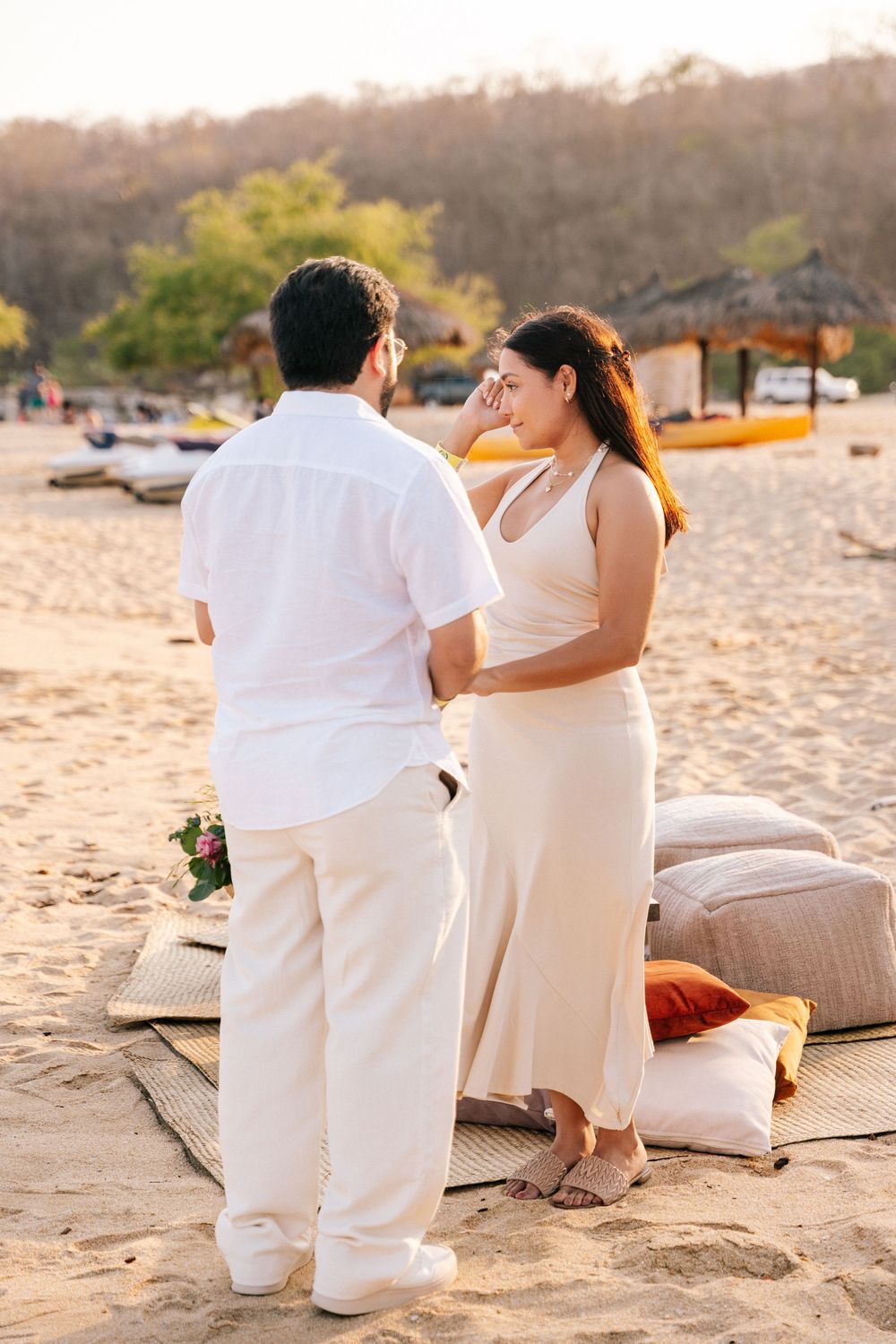 A romantic couple in white outfits shares an intimate moment on a beach at sunset with cushions and decor.