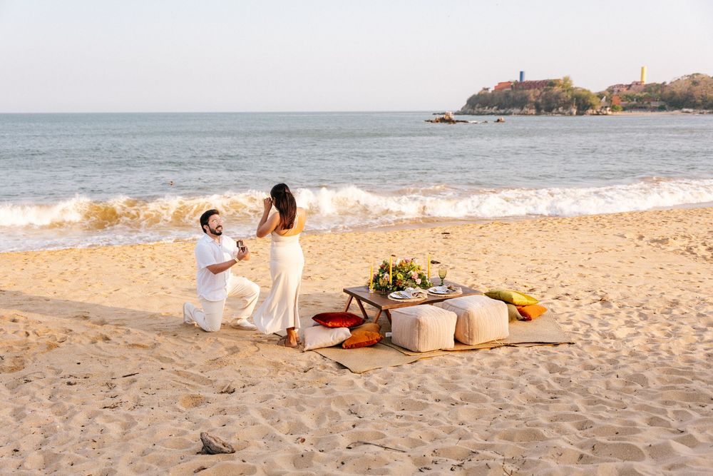Romantic beach picnic setup with blanket, pillows, and decorative elements at sunset along the shoreline.