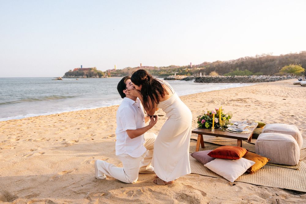 A romantic beach picnic setup with white pillows and blankets on the sand at sunset.