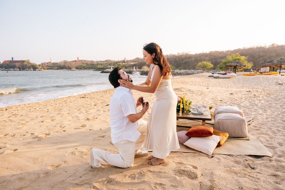 Couple in white outfits sitting together on sandy beach at sunset enjoying romantic moments by the ocean.
