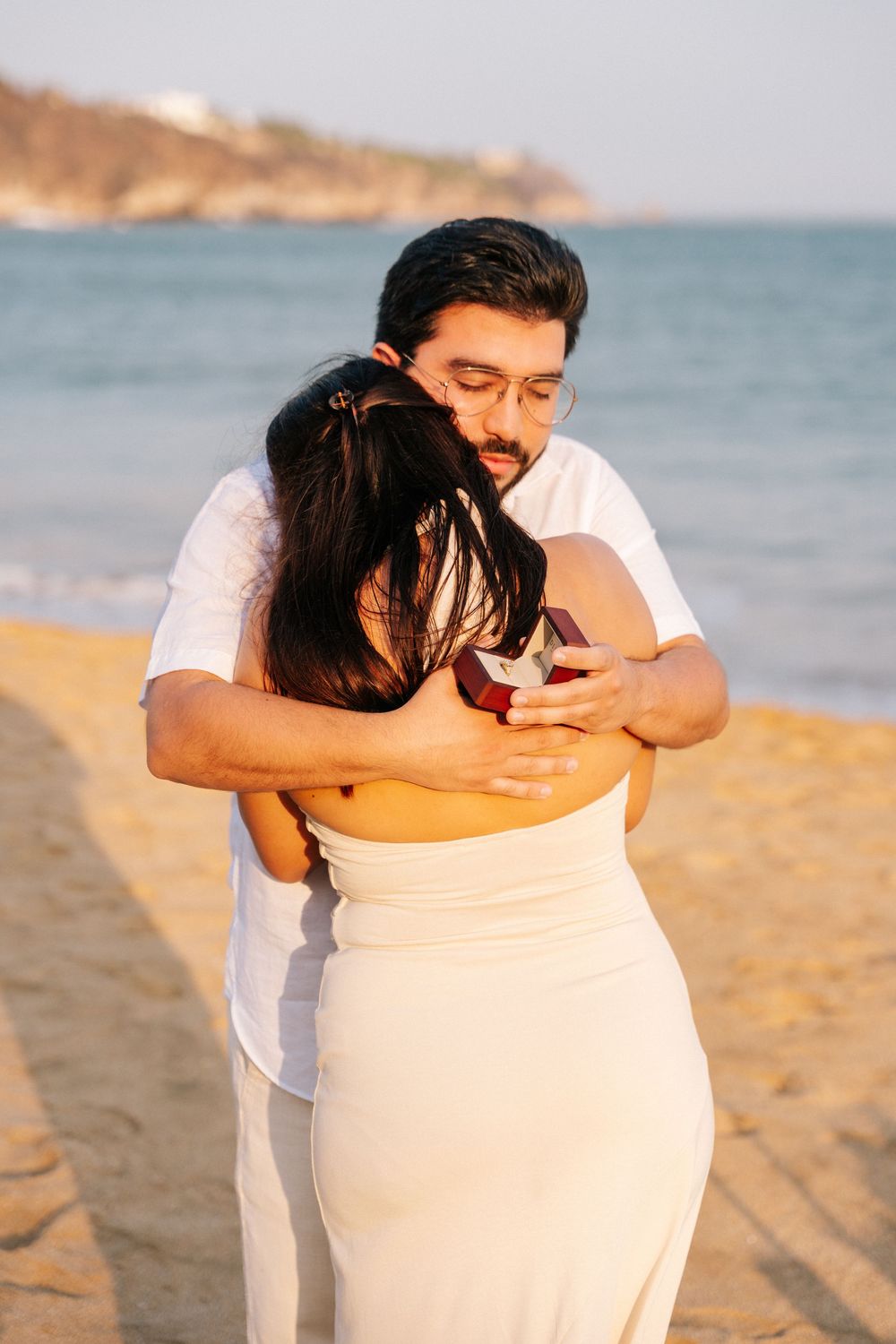 A couple dressed in white embraces tenderly on a sandy beach with ocean cliffs in the background.