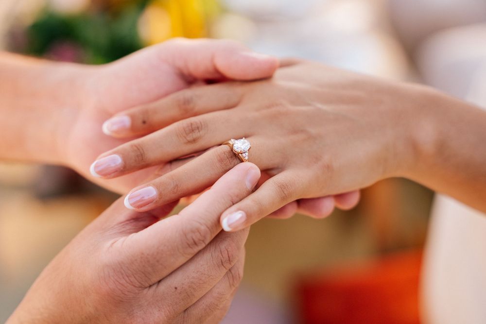 Two hands with a diamond engagement ring exchange a romantic gesture against a soft blurred background.