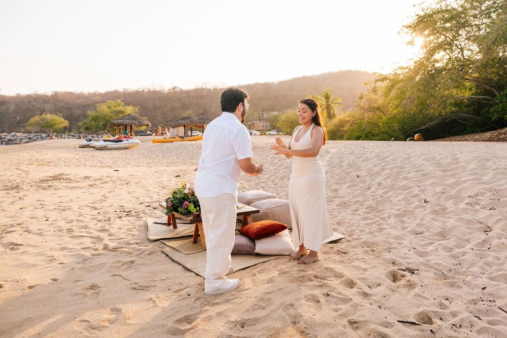 A romantic beach proposal scene at sunset with decorative elements on the sand and boats in the distance.