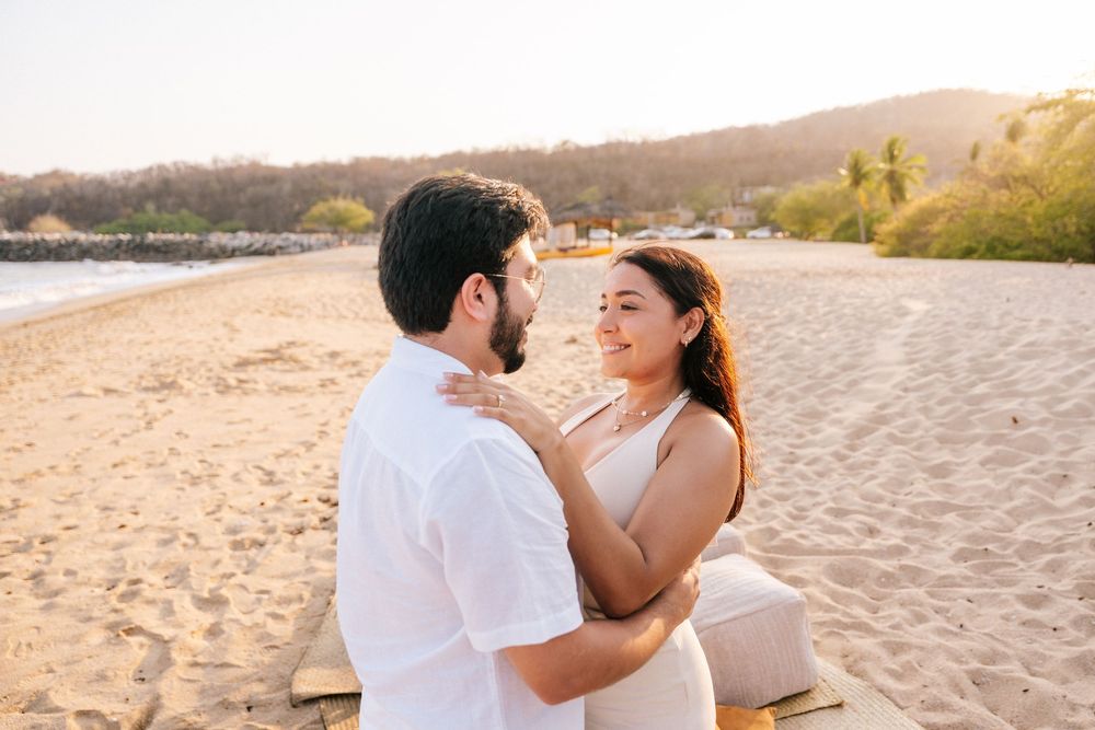 A couple in white outfits embraces romantically on a sandy beach at sunset.