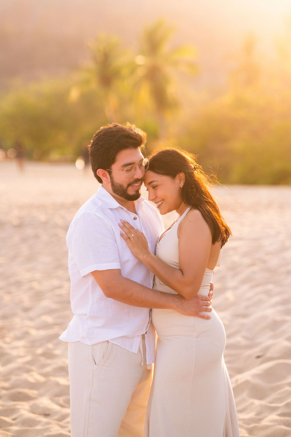 A romantic couple embraces on a sandy beach at sunset wearing white outfits while sharing an intimate moment.