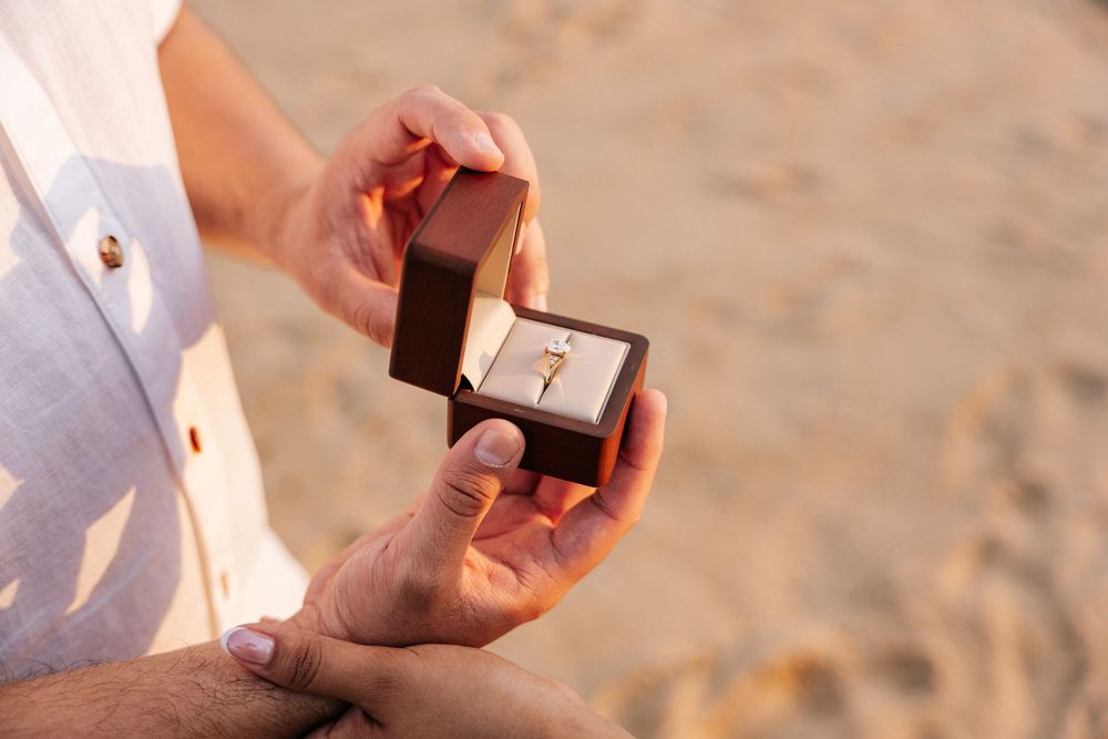 Close-up view of hands exchanging a small ring box during a beach proposal at sunset.