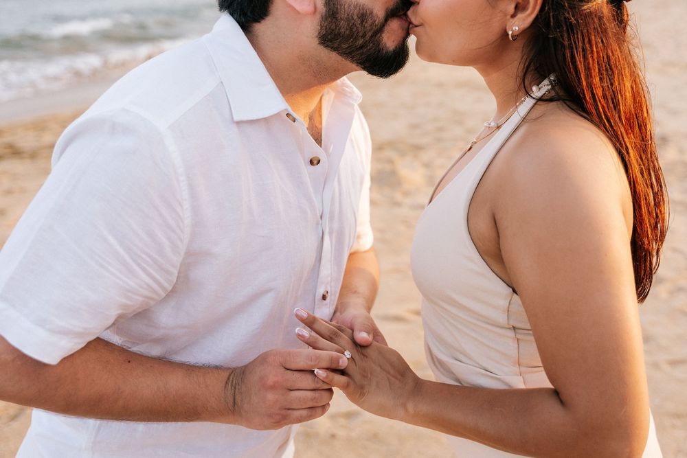 A romantic couple in white outfits shares tender moments on a beach during a photoshoot.