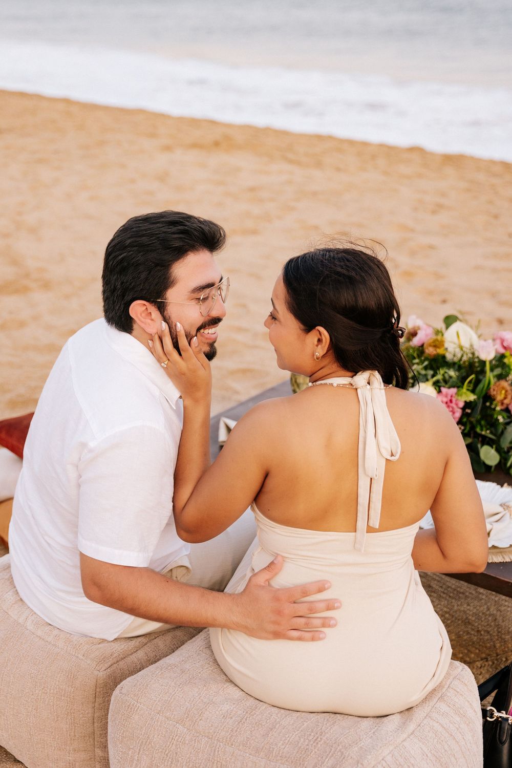 Romantic couple in white and beige outfits share intimate moments while sitting on a beach at sunset.