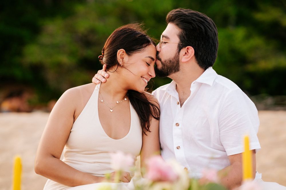 Romantic couple shares tender moments on a beach during sunset photoshoot in white attire.