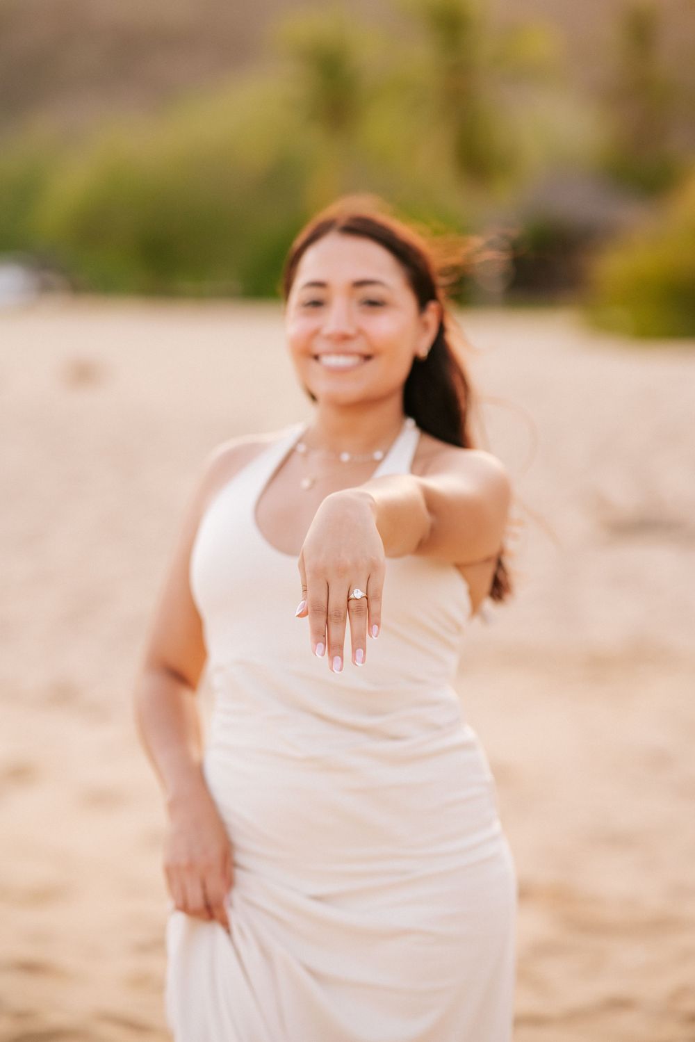 Woman in white dress poses joyfully on sandy beach showing off engagement ring in natural lighting.