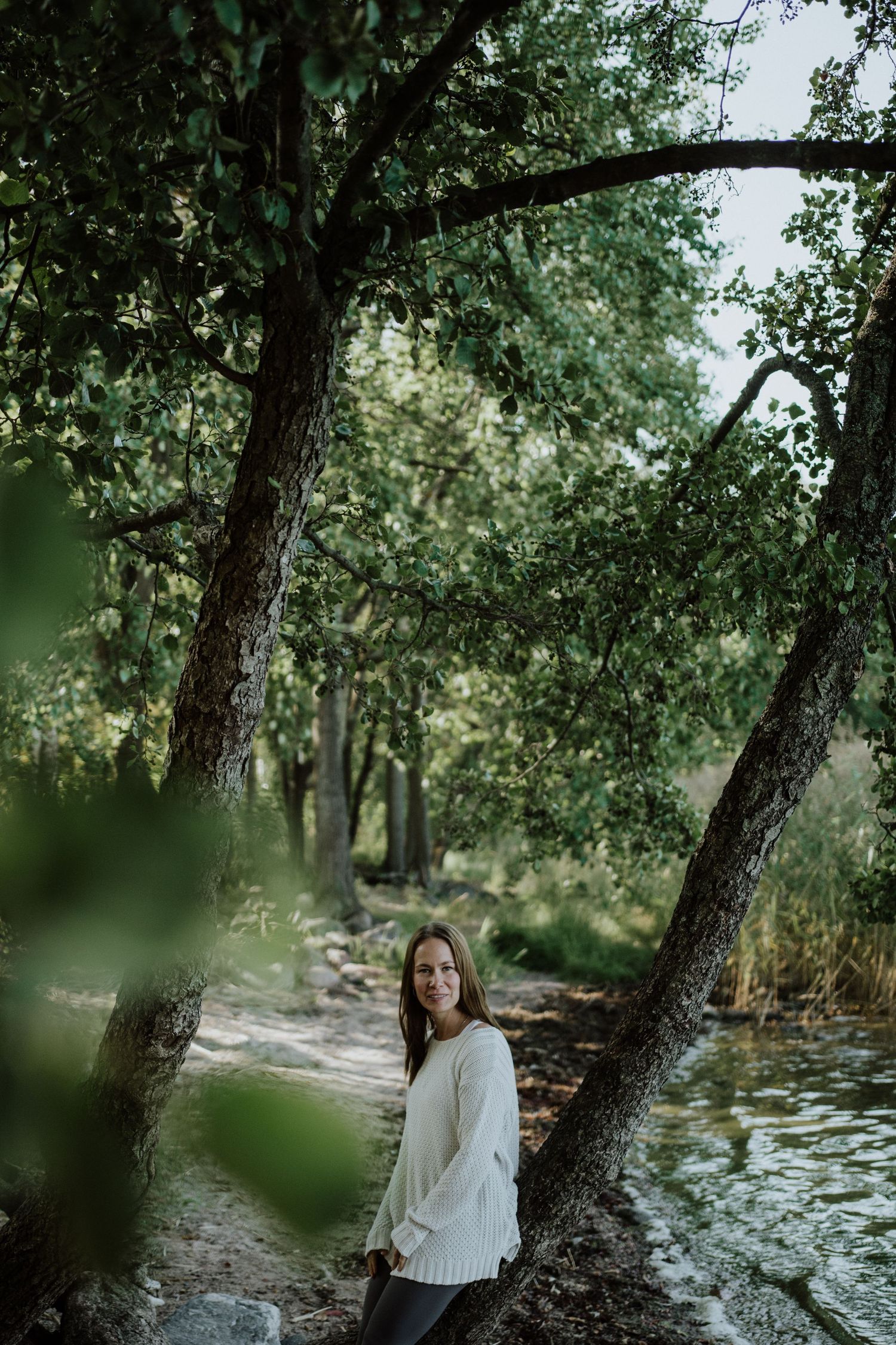 A person in a white sweater stands by a stream in a wooded area on a tranquil autumn day.