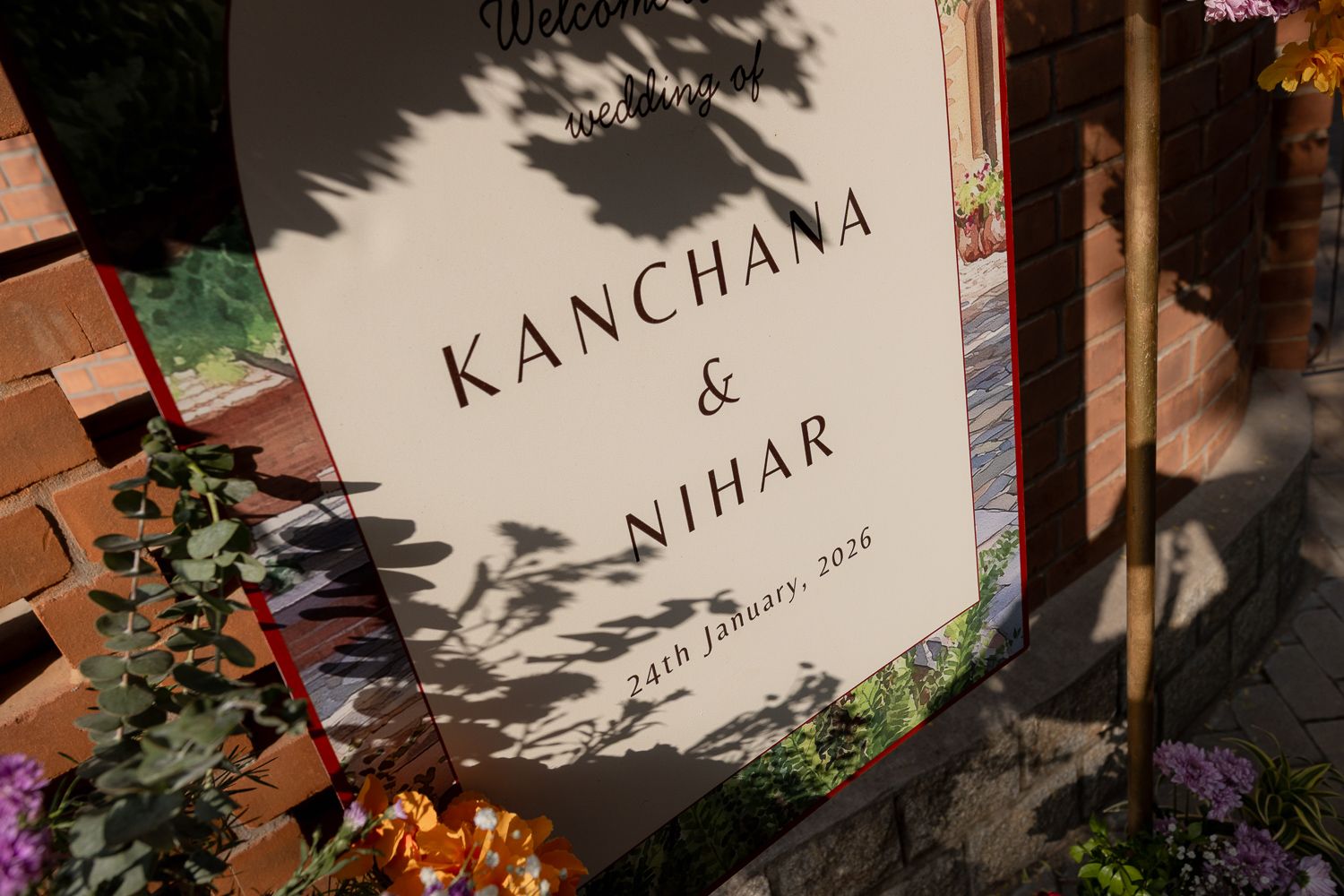 A wedding sign reading 'Kanchana & Nihar' casts shadows on a white surface surrounded by purple flowers.