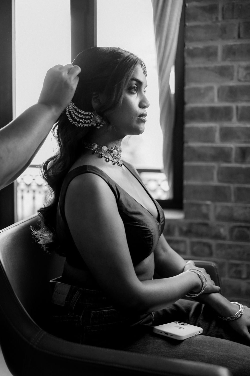 A woman in black sits thoughtfully by a window with exposed brick walls in a dramatic black and white portrait.