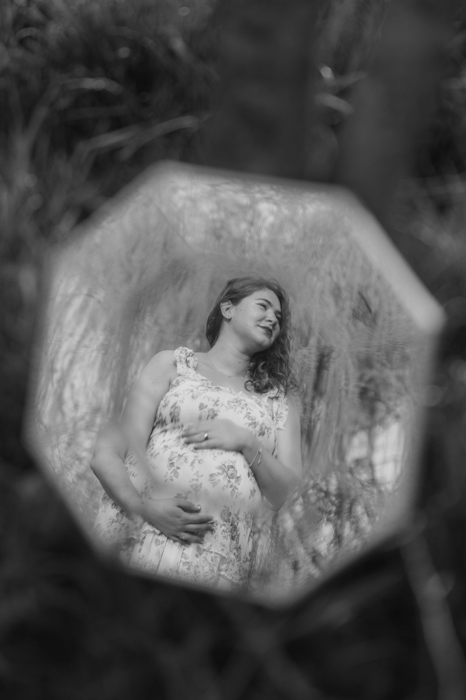 Black and white maternity photo of pregnant woman in floral dress holding belly, captured through octagonal mirror outdoors.