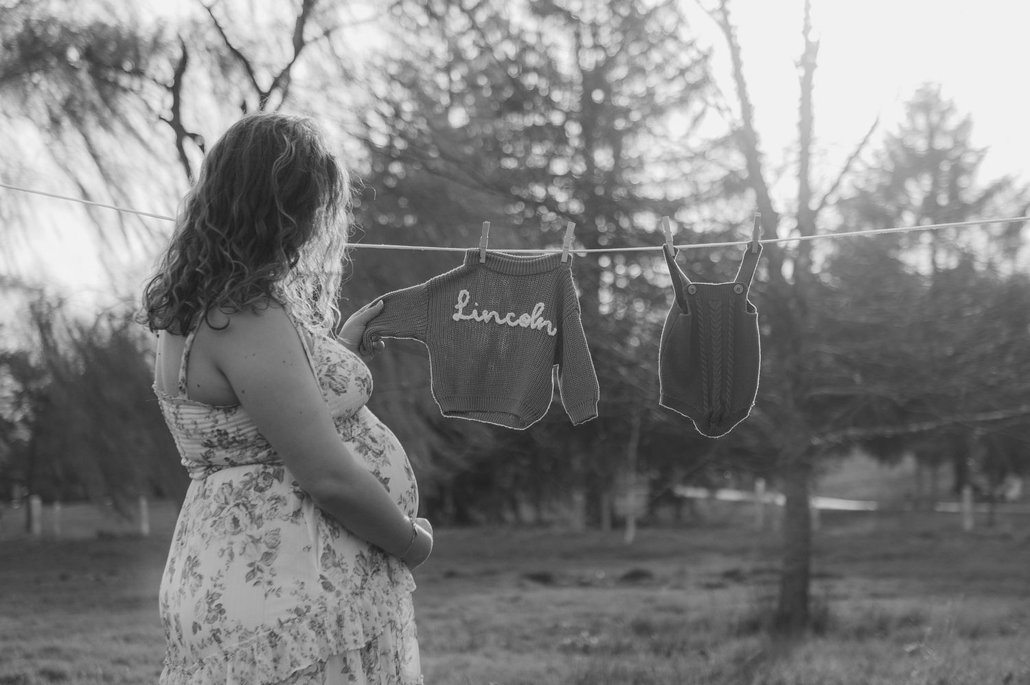 Pregnant woman in floral dress stands beside clothesline with baby clothes spelling Lincoln, black and white maternity photo.