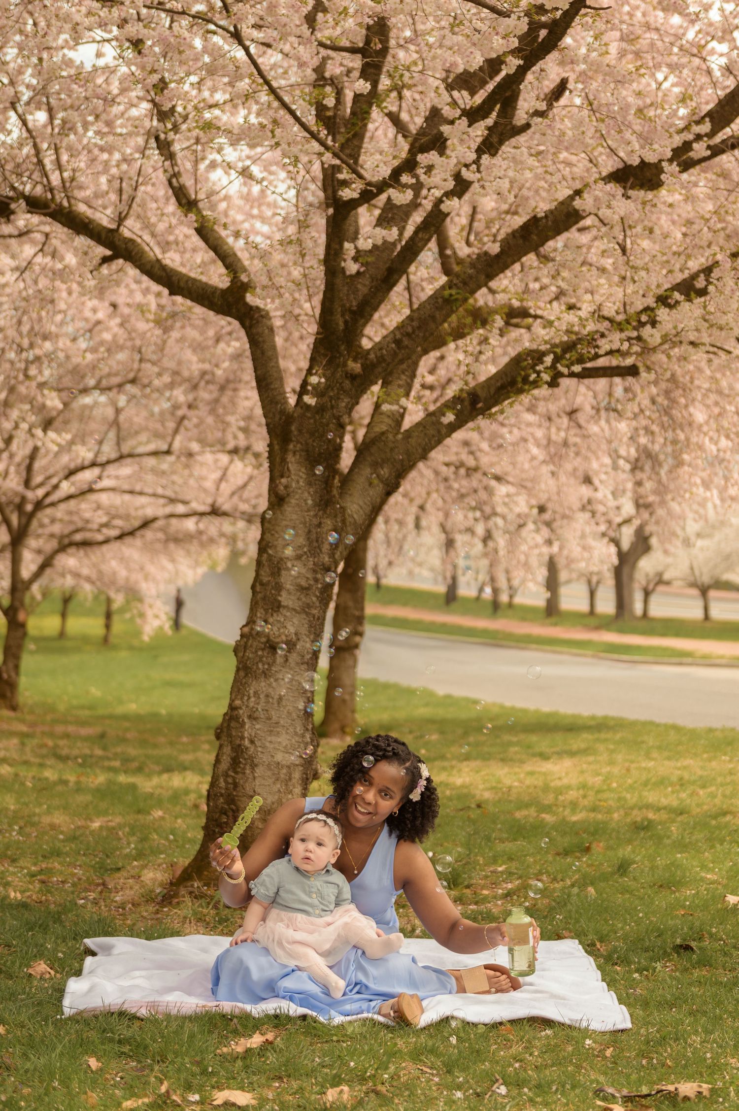 Mother and baby daughter enjoy a spring picnic under blooming cherry blossom trees on a white blanket in a lush park.