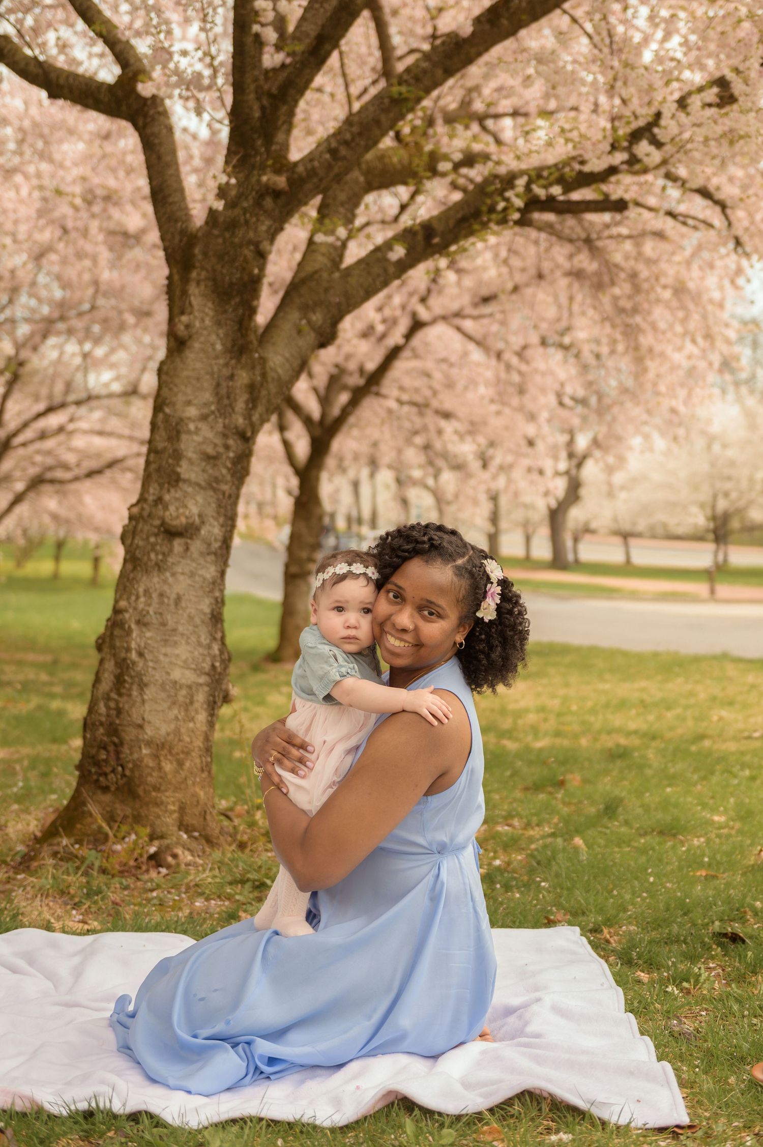 A mother in a blue dress sits on a blanket holding her baby under blooming cherry blossom trees in a park.