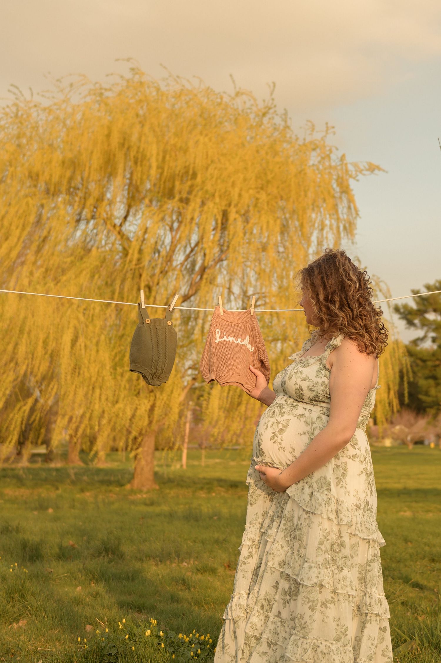 Pregnant woman in floral dress holding belly near clothesline with baby clothes, golden willow tree backdrop at sunset.