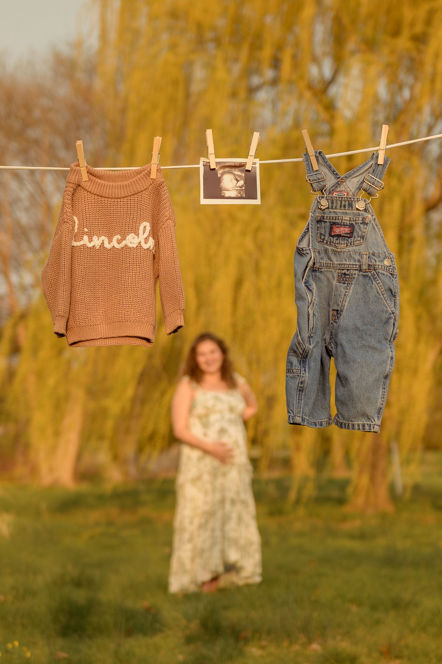 Maternity photo with baby clothes and ultrasound on clothesline, pregnant woman in floral dress standing behind near willow tree.