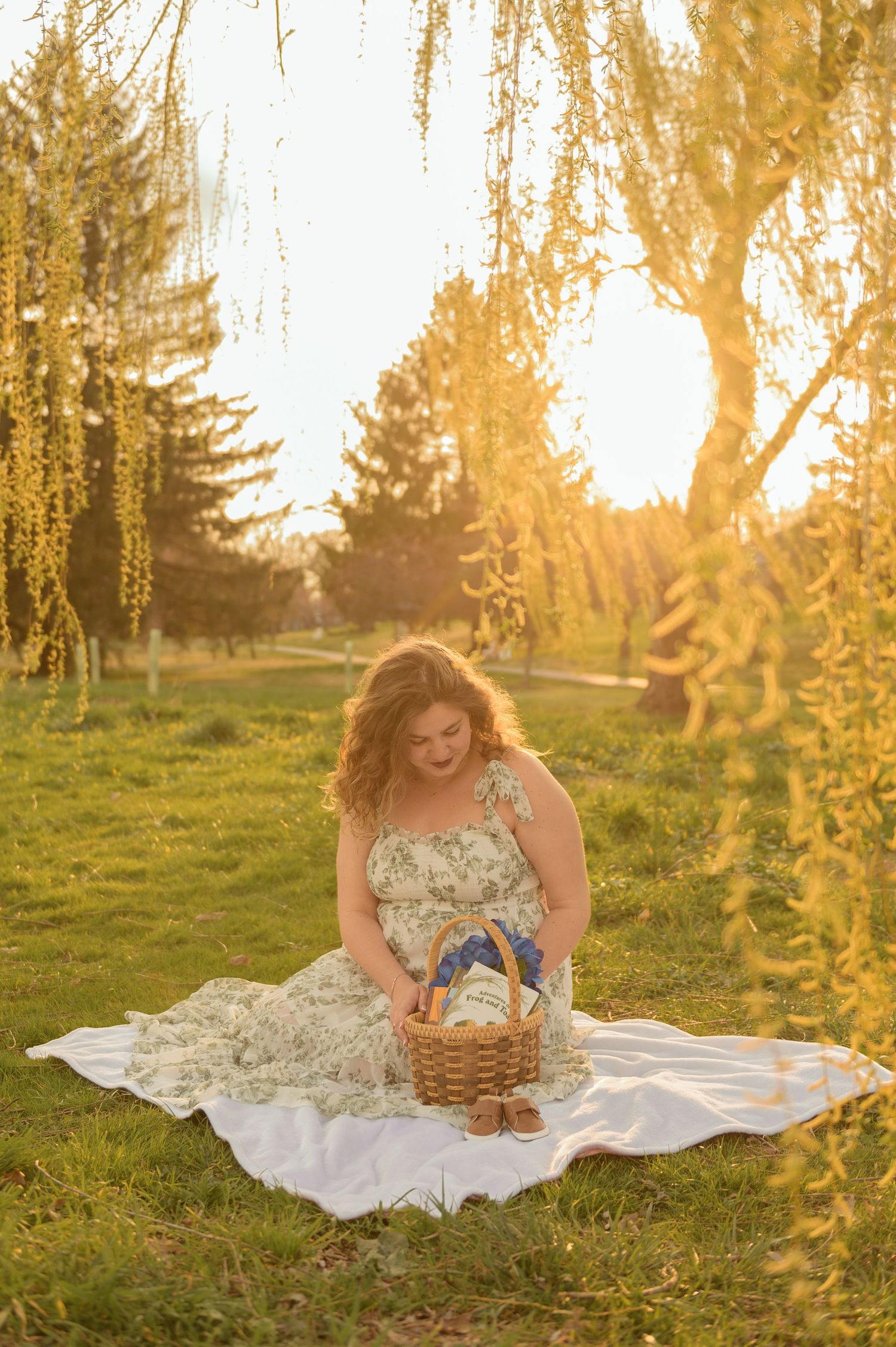 Woman in floral dress sits on white blanket with basket under weeping willow tree at golden hour sunset.