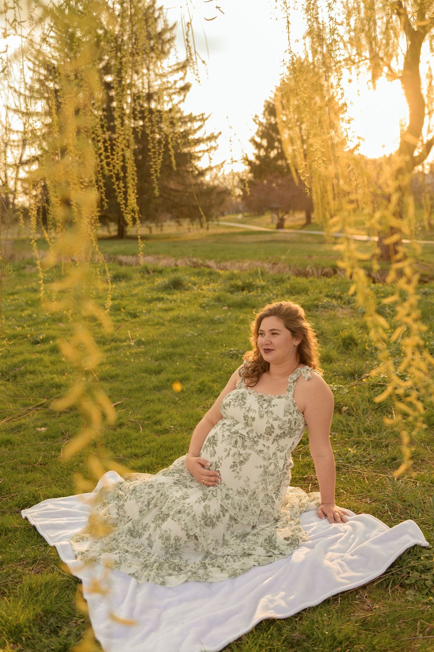 Pregnant woman in floral dress sitting on white blanket outdoors at golden hour, surrounded by weeping willow branches.