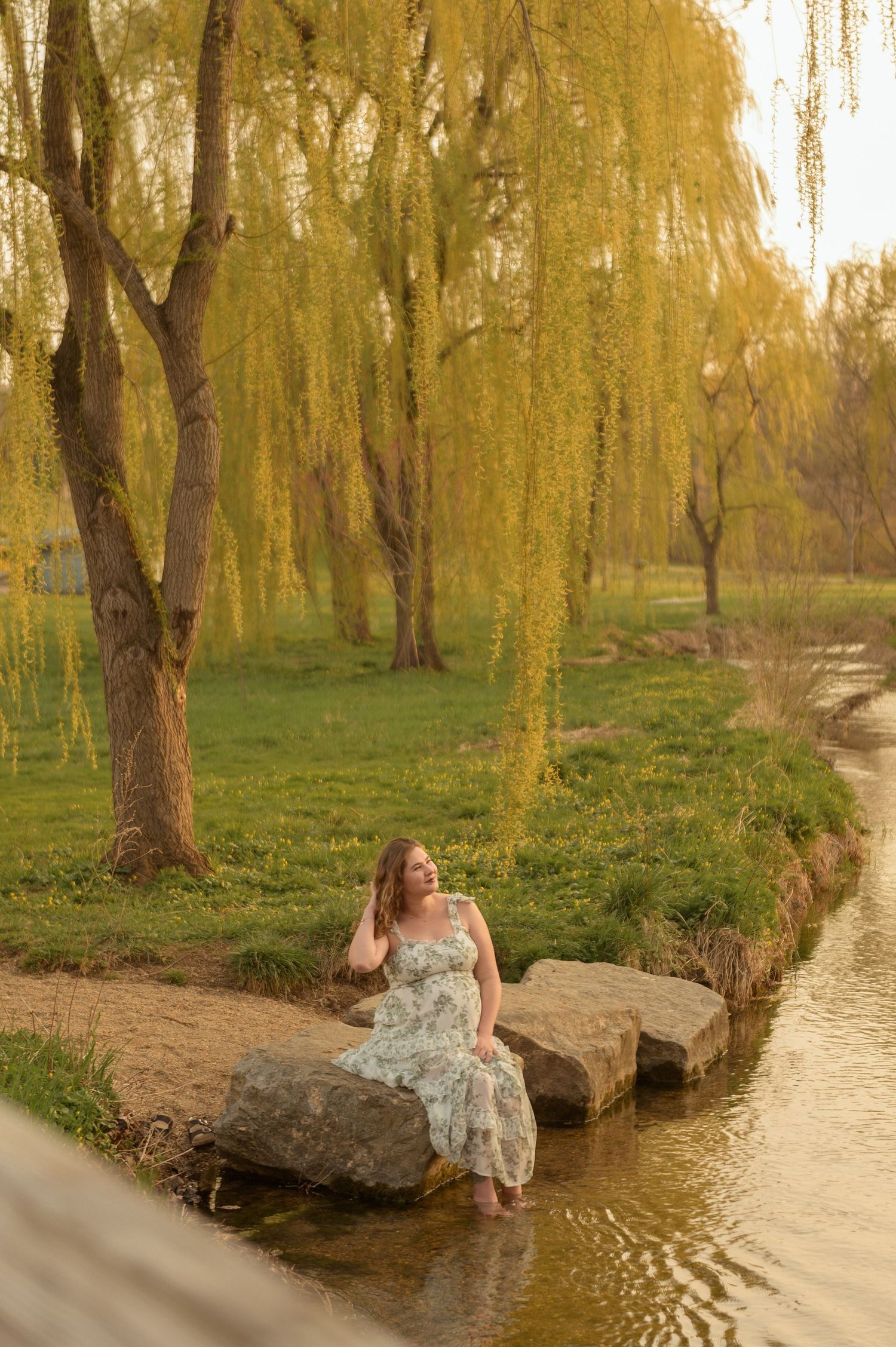 Woman in floral dress sitting on rocks by a stream under weeping willow trees in golden hour light.