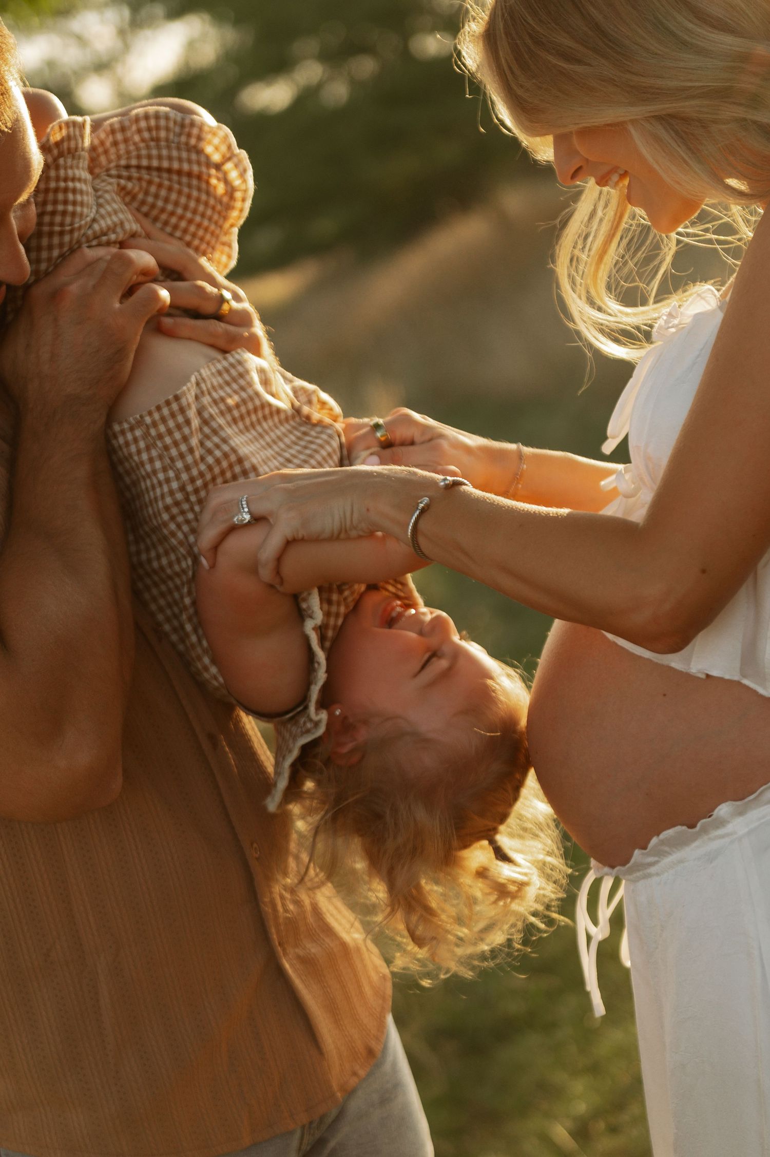 A joyful family moment outdoors in golden light, with parents playfully holding a laughing toddler upside down.