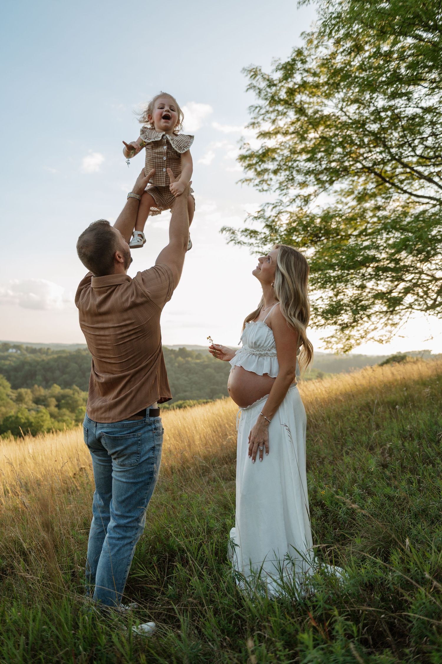 Pregnant woman in white dress watches husband lift toddler in sunny meadow during outdoor maternity photo session.