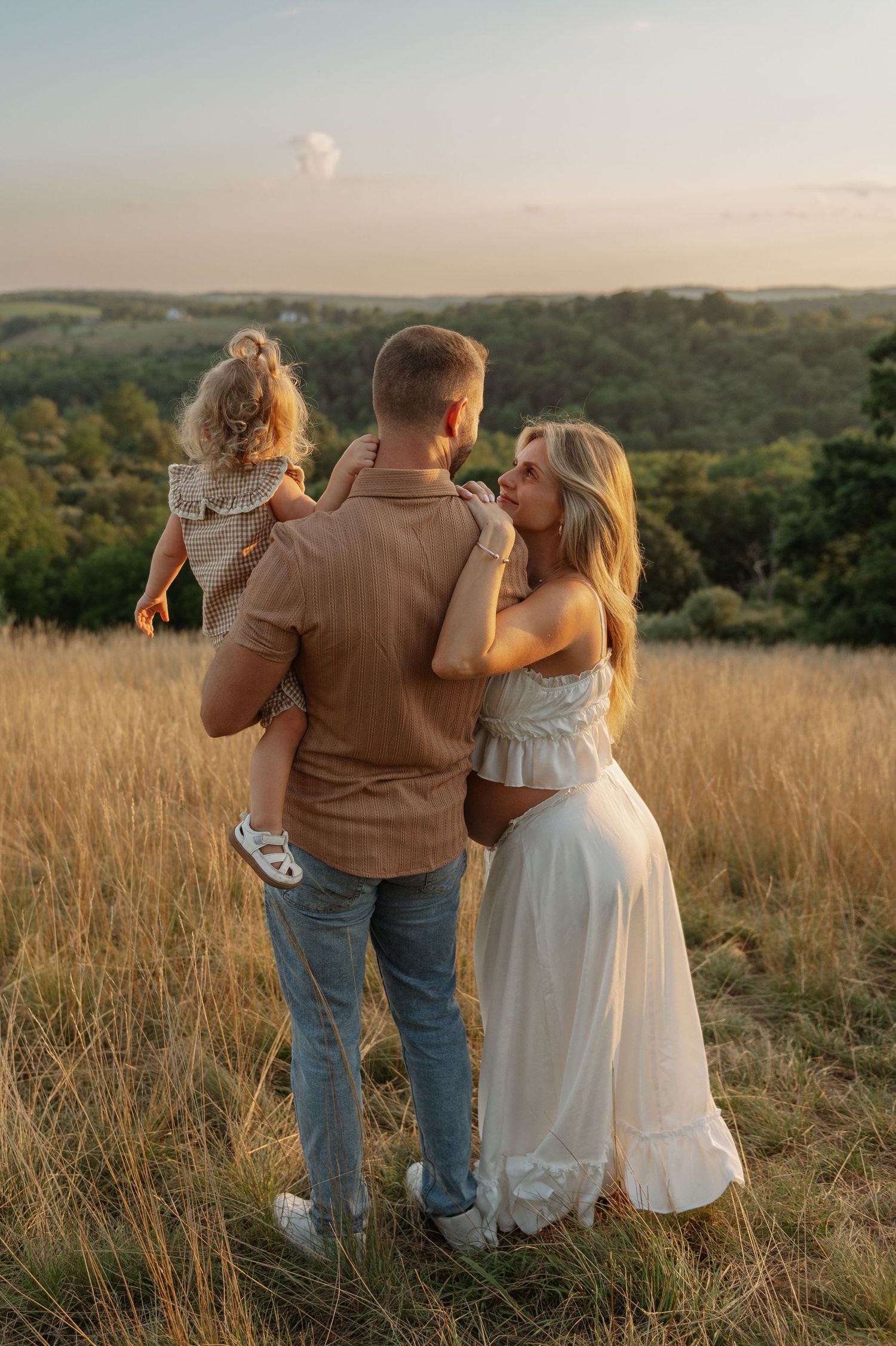A family of three stands in a golden field at sunset, with a man holding a toddler and a pregnant woman in a white dress.