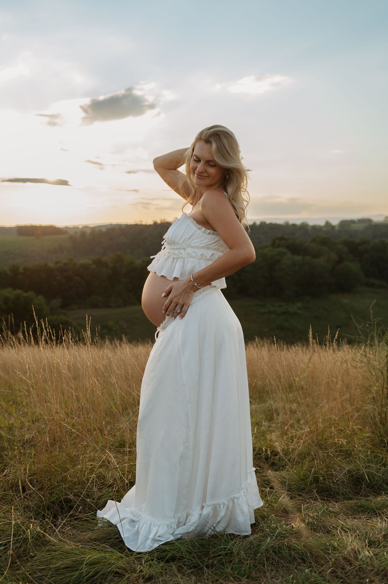 Pregnant woman in white two-piece dress posing in golden field at sunset, smiling with hand behind head.