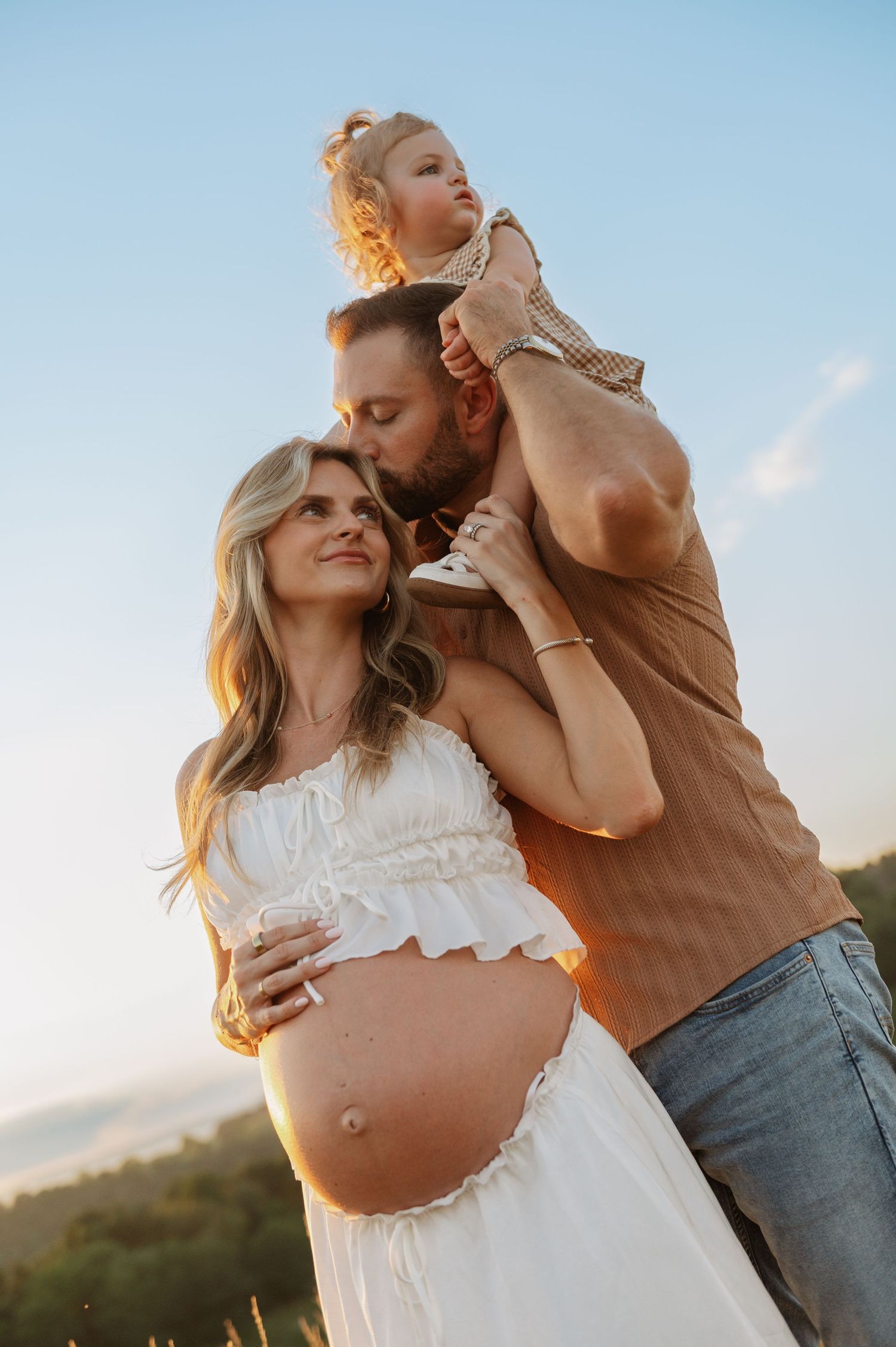 Pregnant woman in white dress with partner and toddler on shoulders during golden hour outdoor maternity photoshoot.