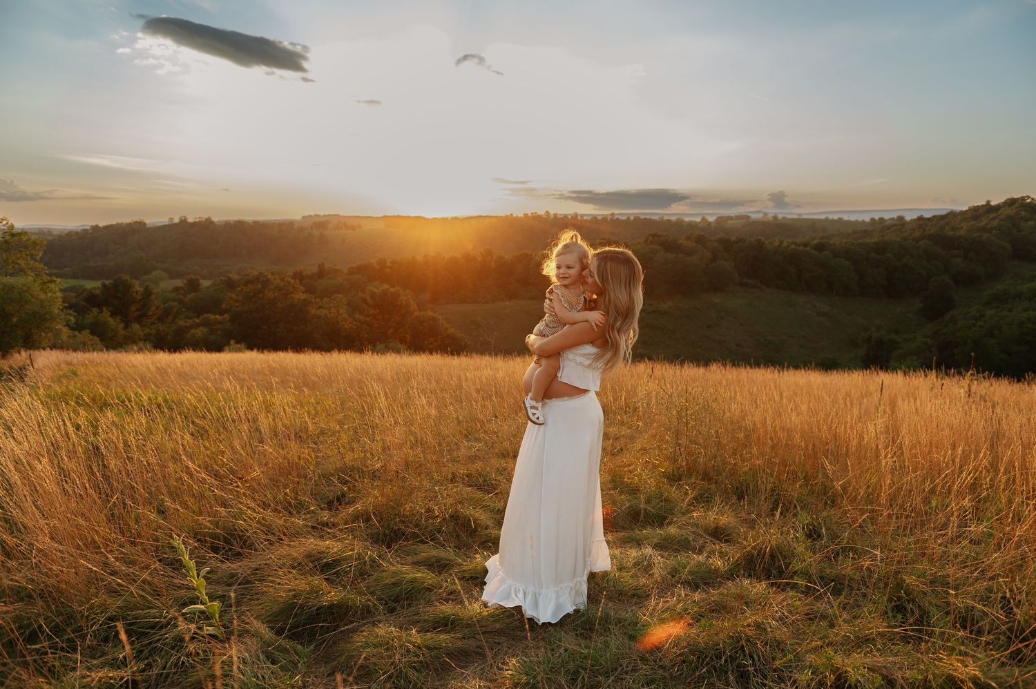 Woman in white dress holding a child in a golden wheat field at sunset with rolling hills in the background.