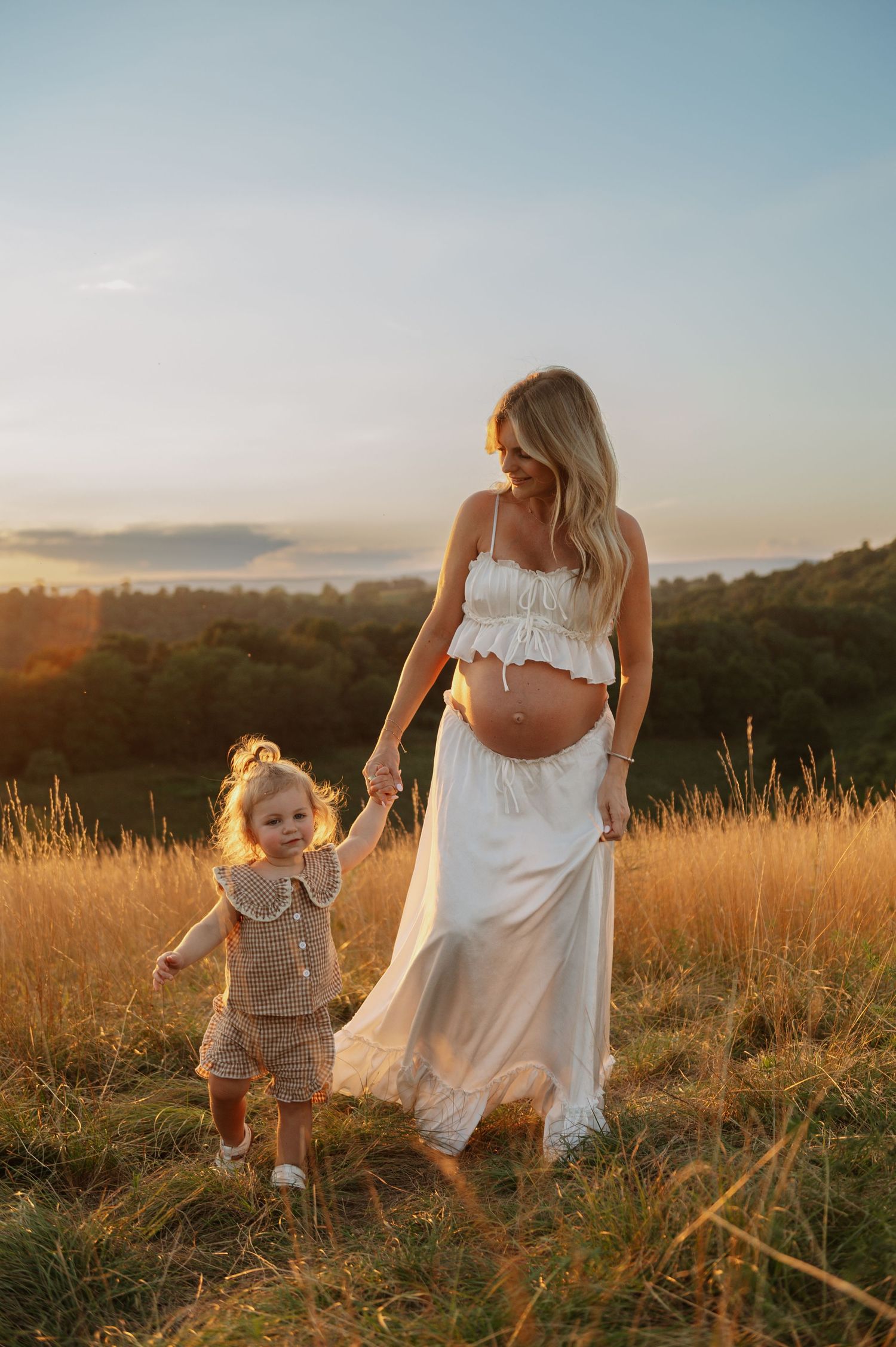 Pregnant woman in white crop top and skirt holds toddler's hand in golden field at sunset.