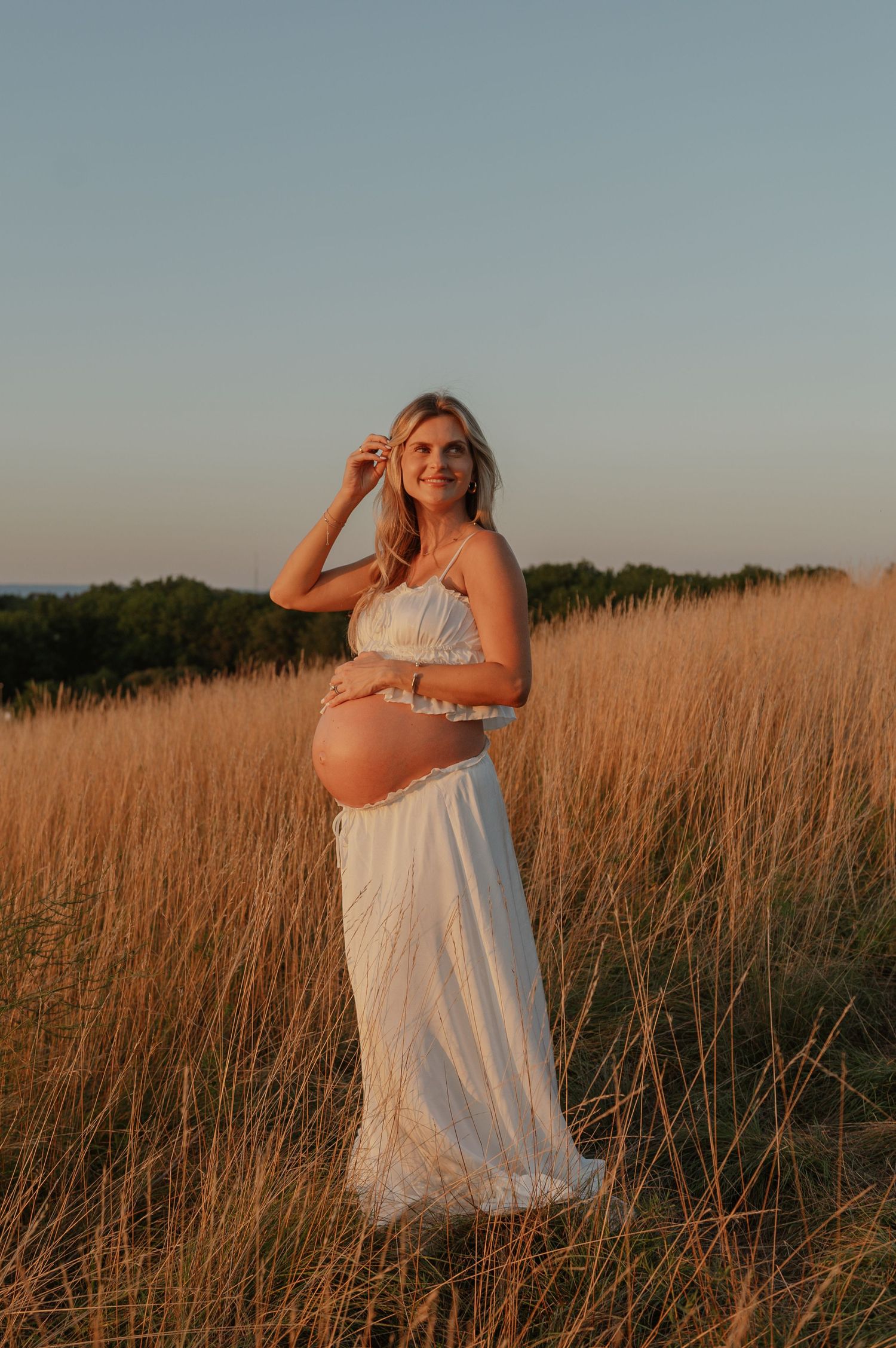 Pregnant woman in white dress standing in golden field at sunset, cradling baby bump with one hand raised to hair.