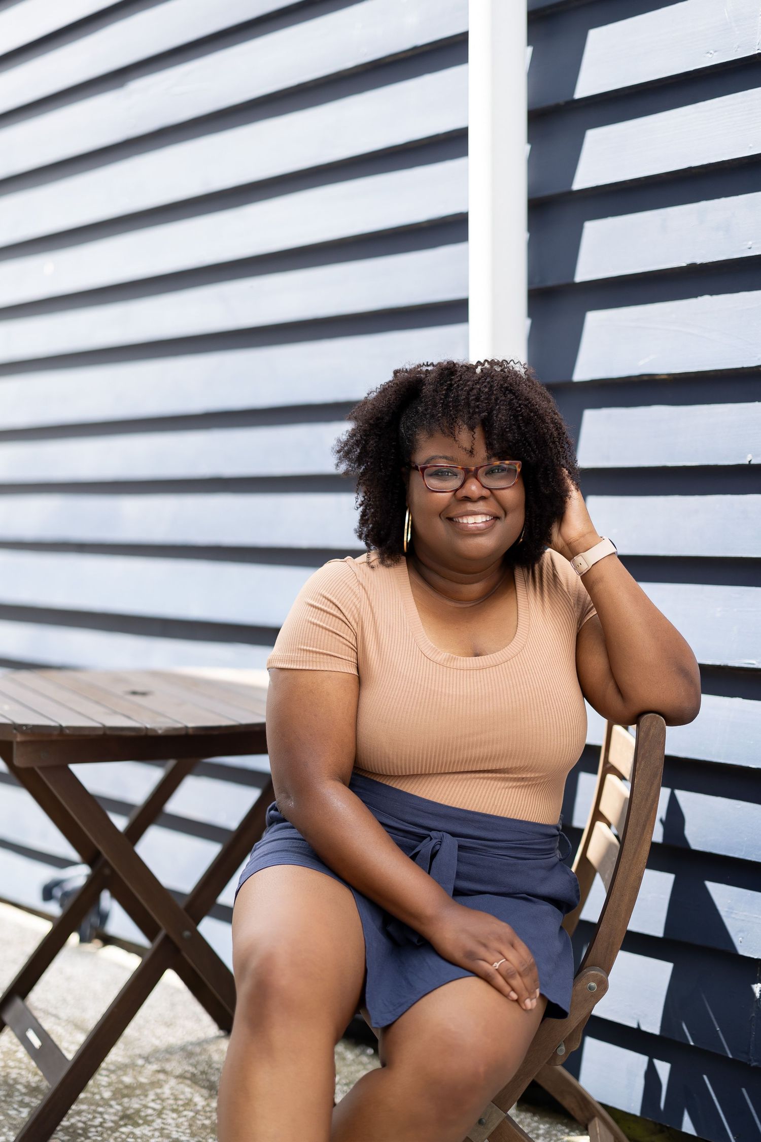 A smiling plus-size Black woman with glasses and natural hair sits casually in a wooden chair against a blue siding wall.