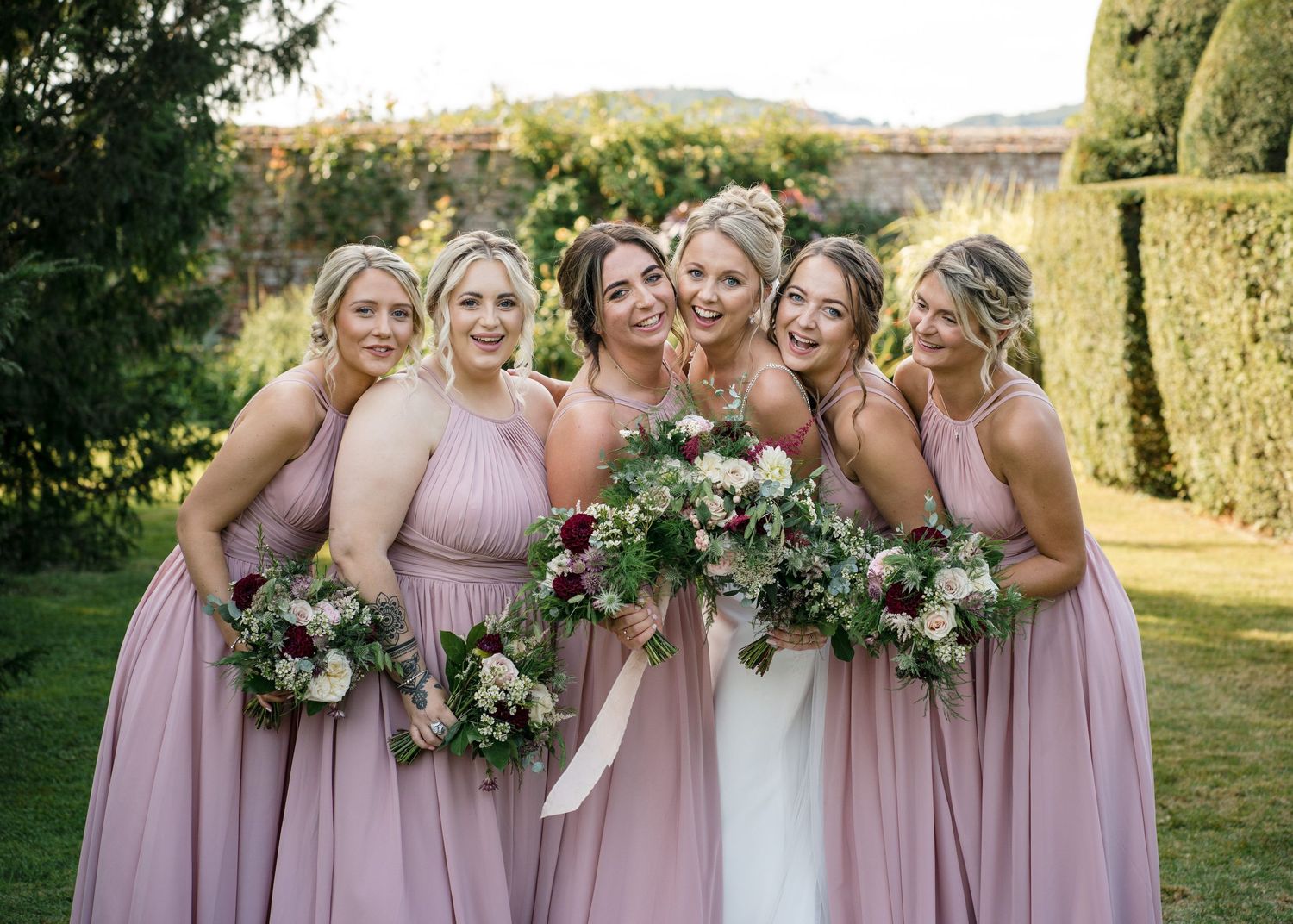 Bridesmaids in dusty rose dresses pose together with floral bouquets during an outdoor wedding celebration.
