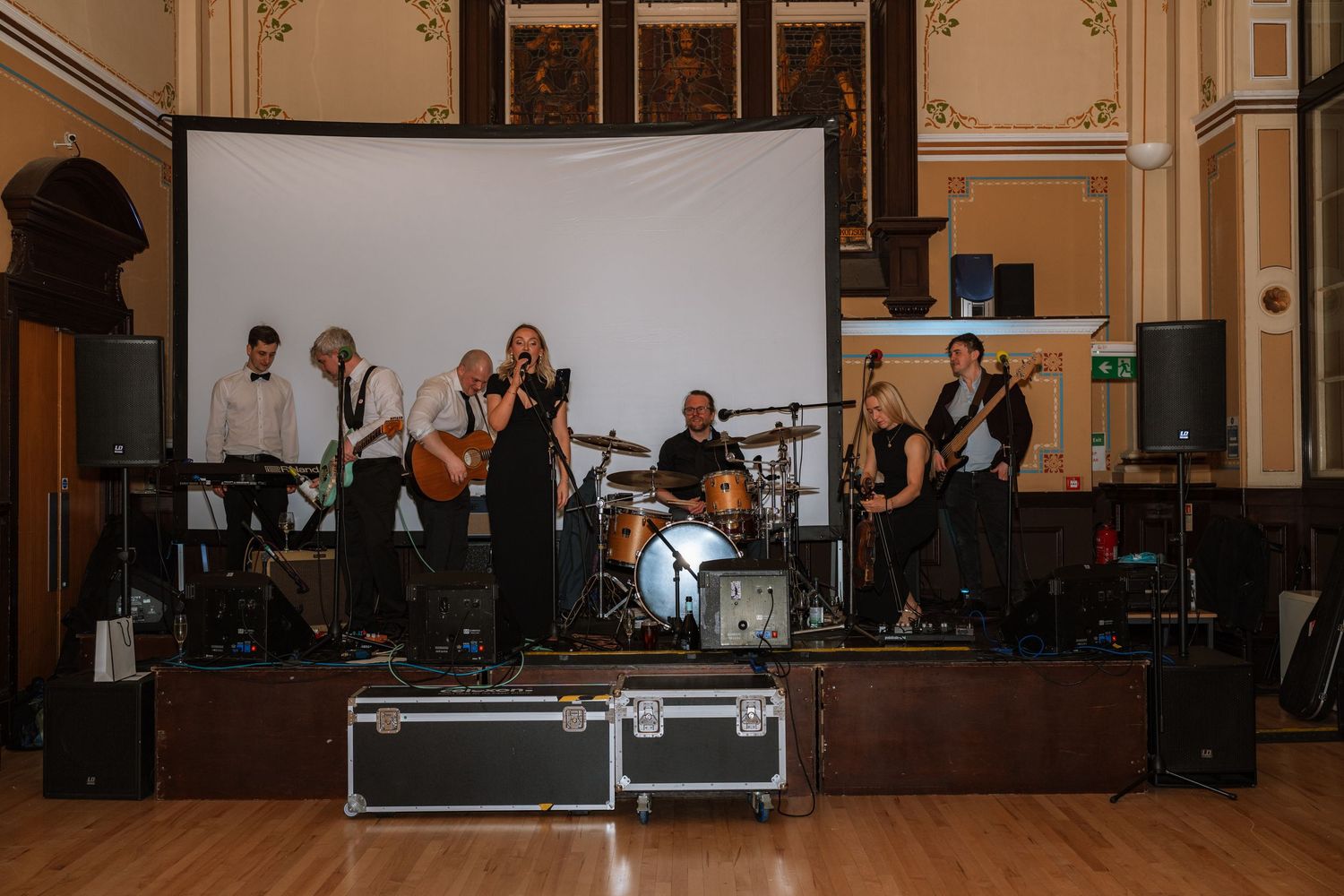 Musicians perform on stage during a live concert in an elegant ballroom with ornate architectural details.