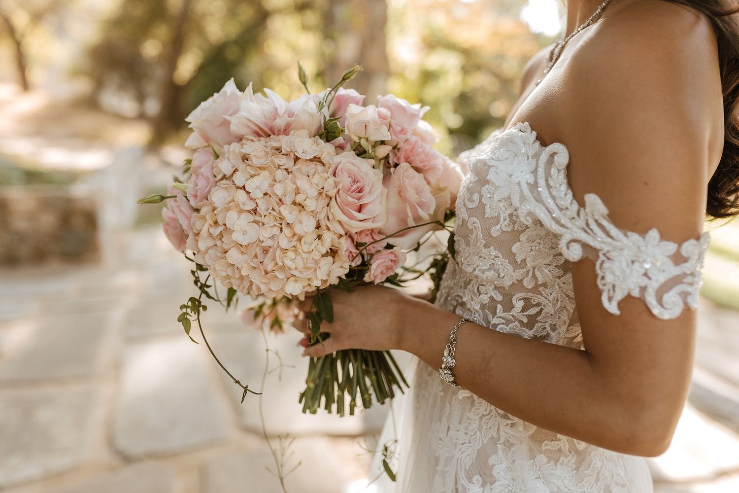 Bride in lace off-shoulder wedding gown holding elegant blush pink hydrangea and peony bouquet outdoors.